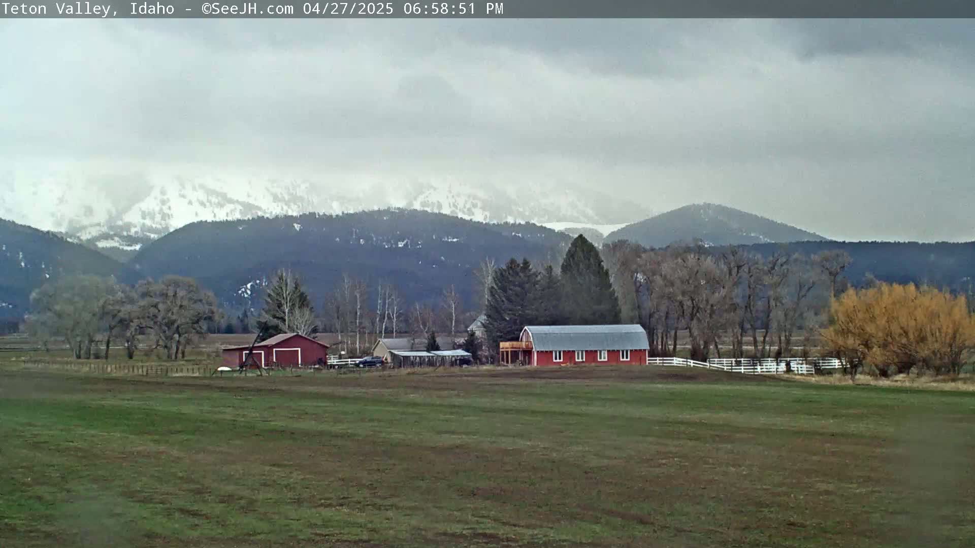 A snow-capped mountain range is visible in the distance behind a flat, grassy field with a red barn and other farm buildings.  The sky is overcast.