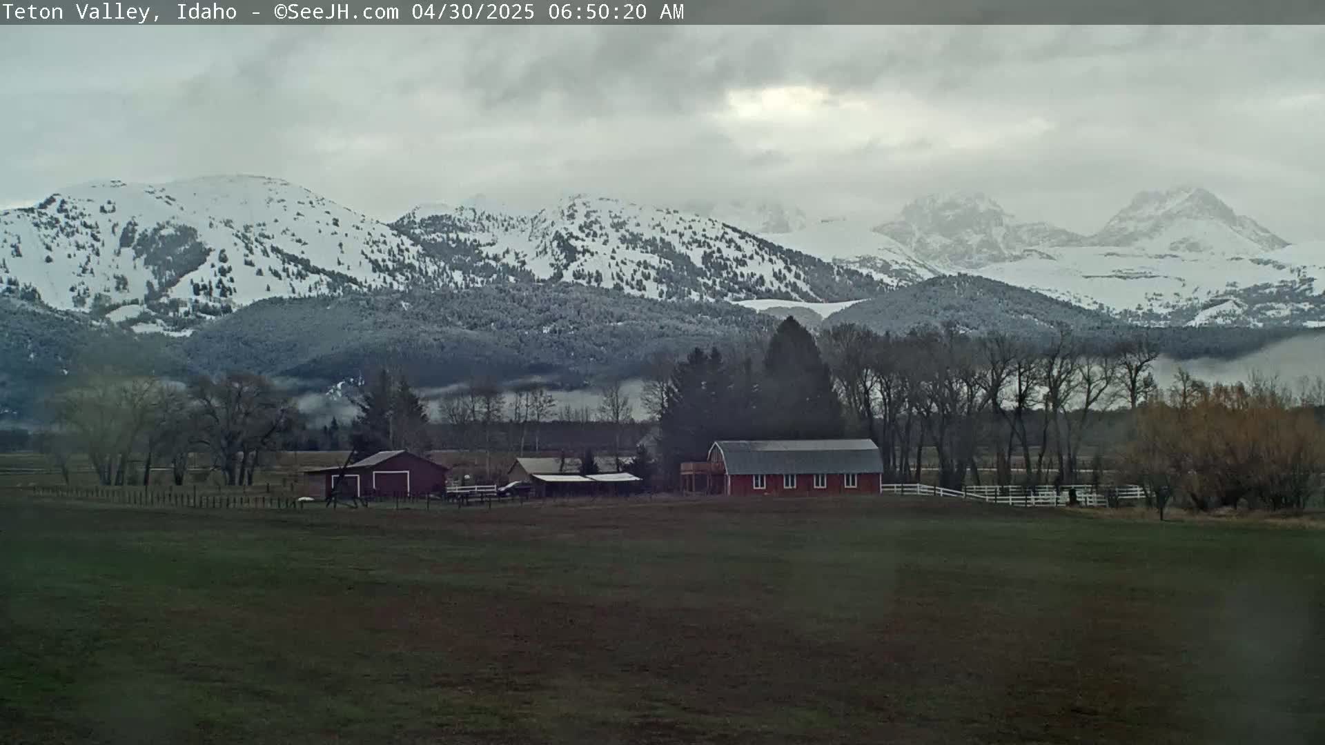 A red barn sits on a grassy field in front of snow-capped mountains under a cloudy sky.