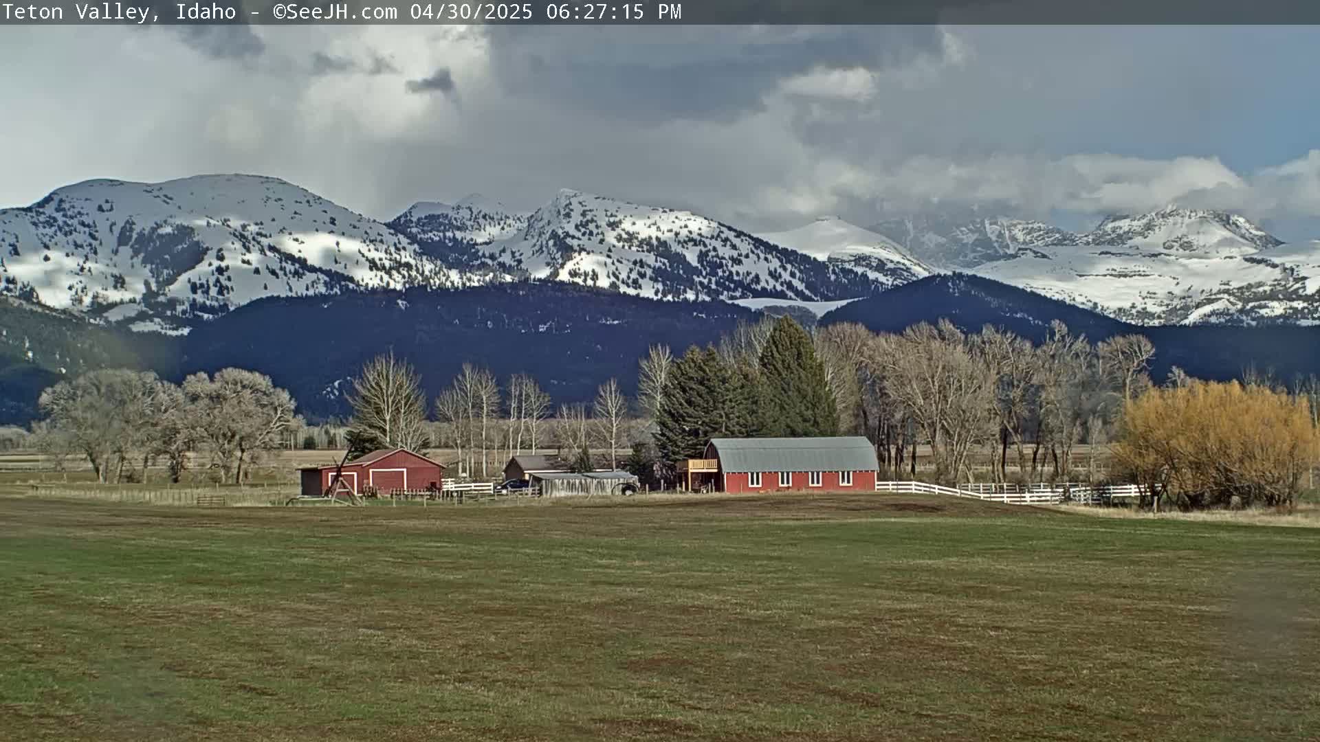 A red barn sits on a grassy field in front of snow-capped mountains under a partly cloudy sky.