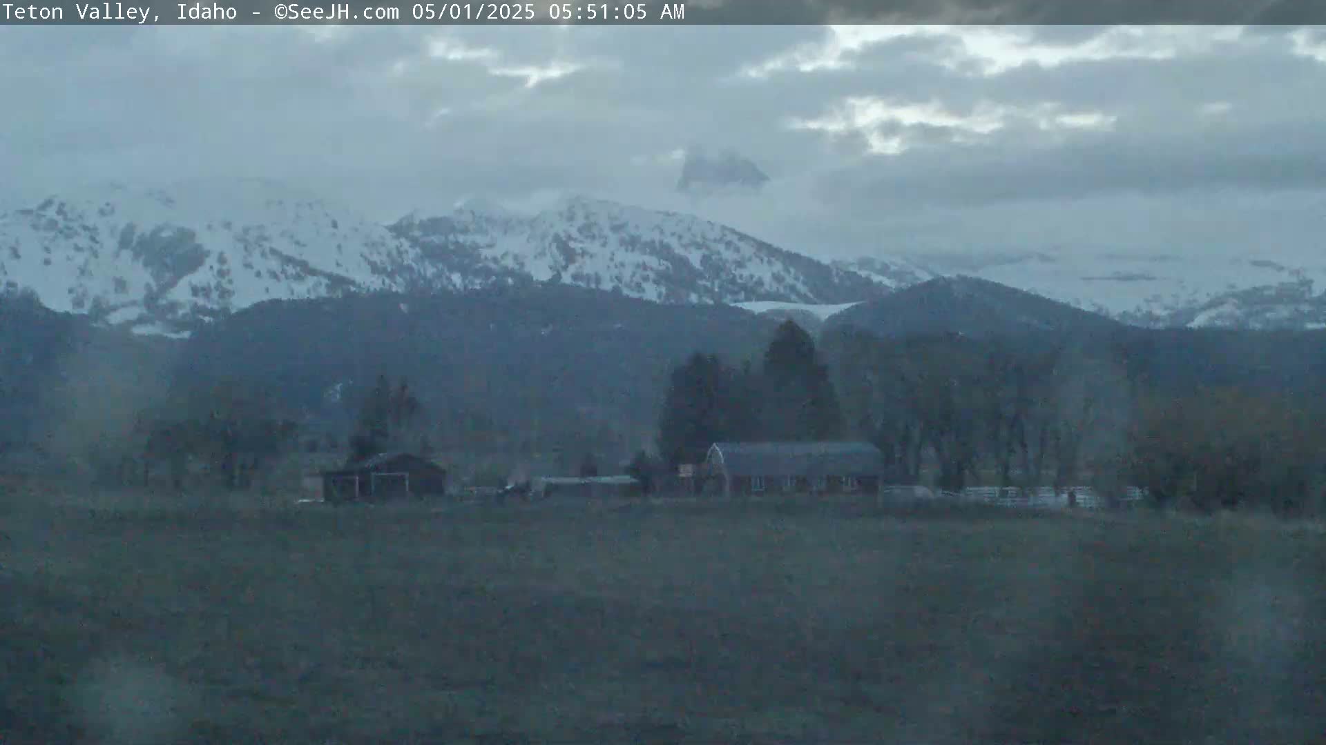 A snow-dusted mountain range is visible in the distance under an overcast sky, with a few buildings and trees in a field in the foreground.
