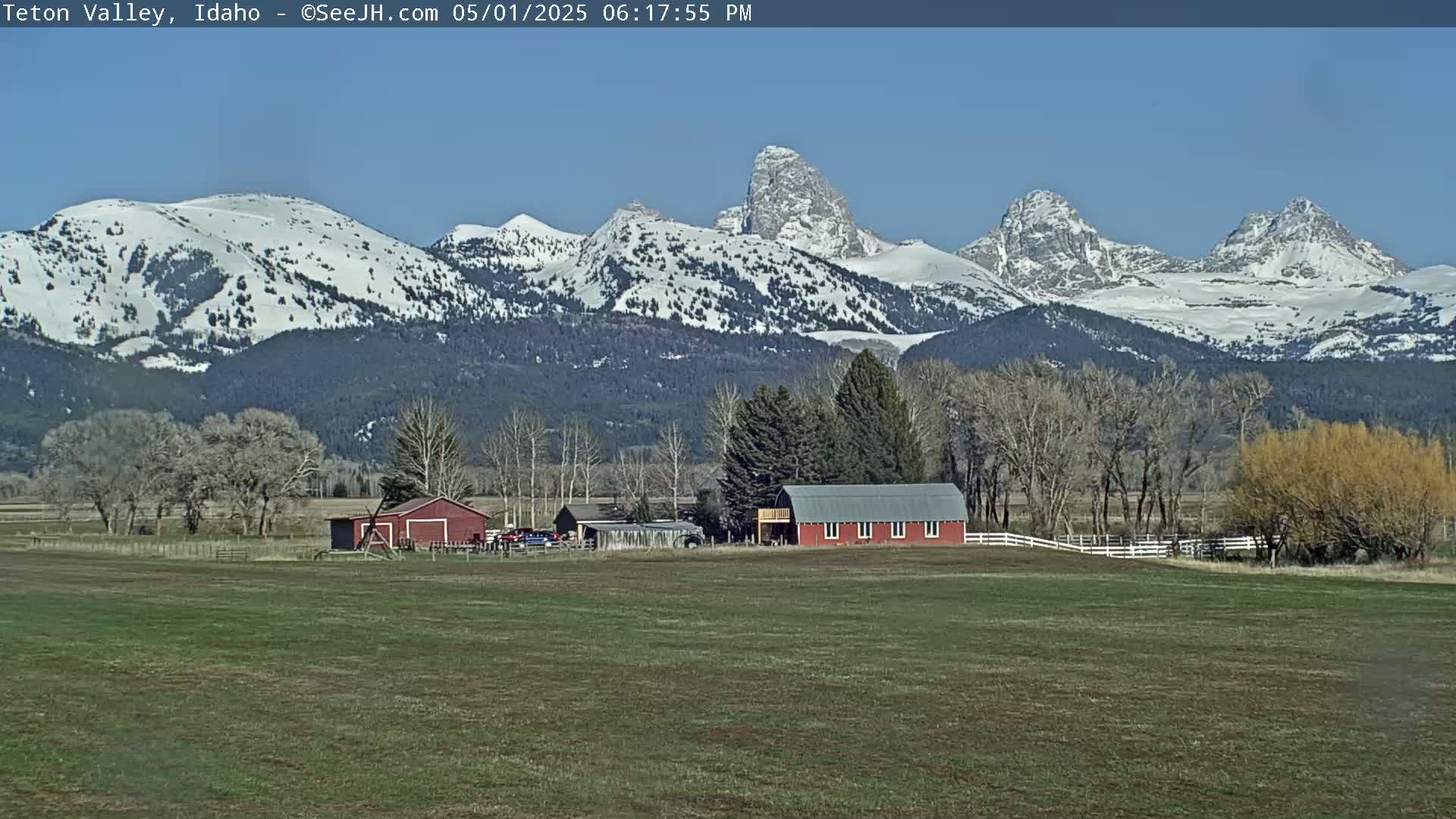 A red barn sits on a grassy field in front of snow-capped mountains under a clear blue sky.