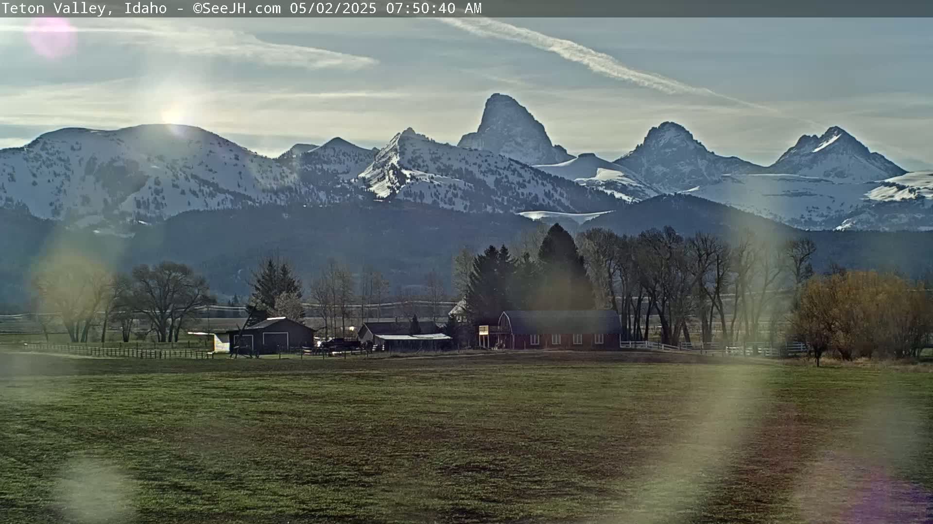 A farm with several buildings sits in a grassy field in front of snow-capped mountains under a partly cloudy sky.