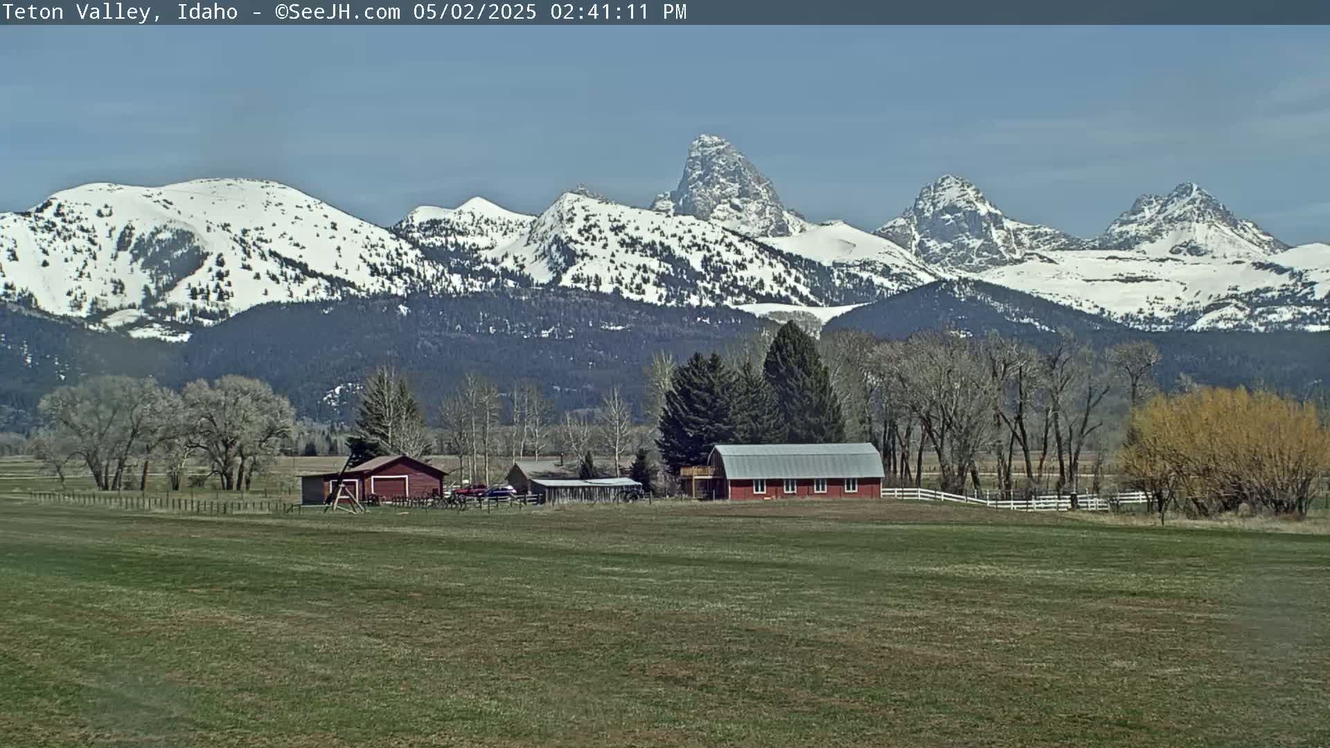 A snow-capped mountain range rises behind a flat field with a red barn and outbuildings under a clear, bright sky.