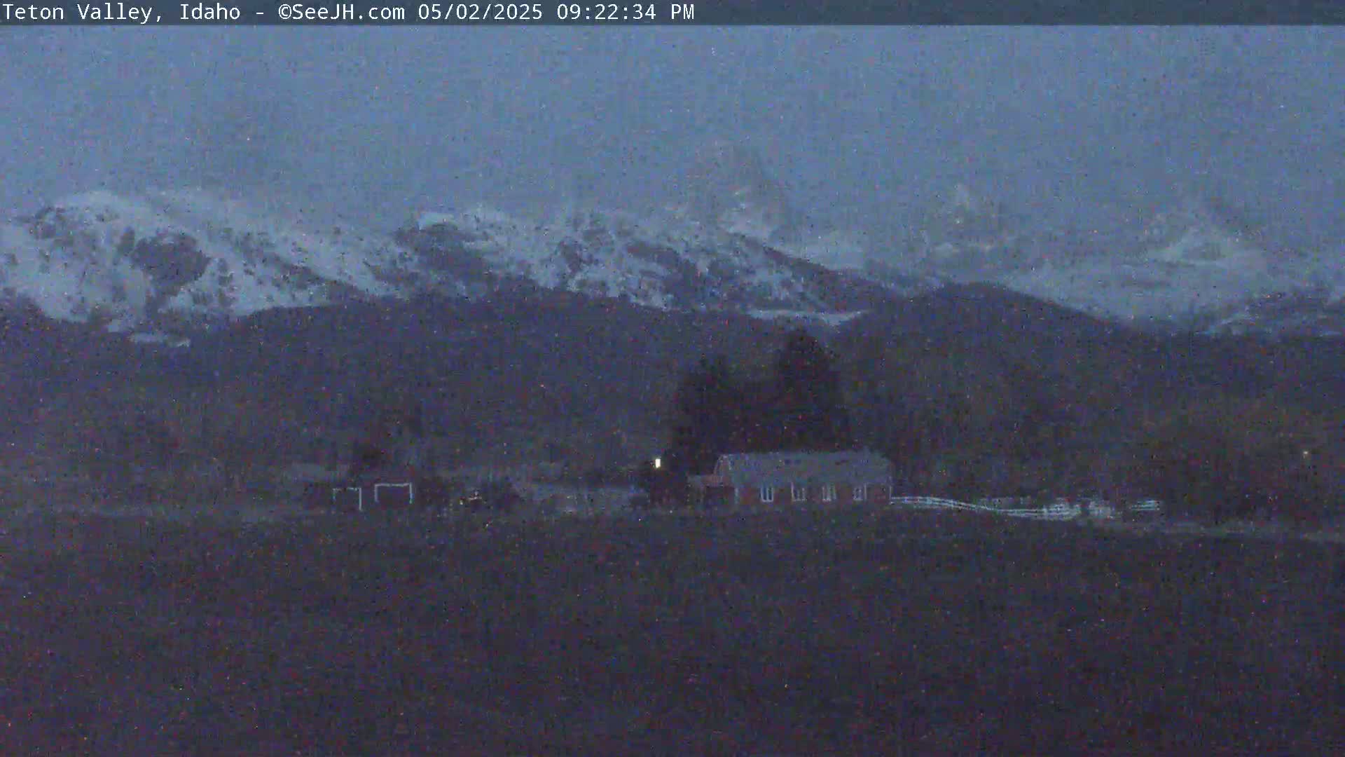 A snow-dusted mountain range is visible in the distance, overlooking a rural landscape with a few buildings under a dark, possibly snowy sky.