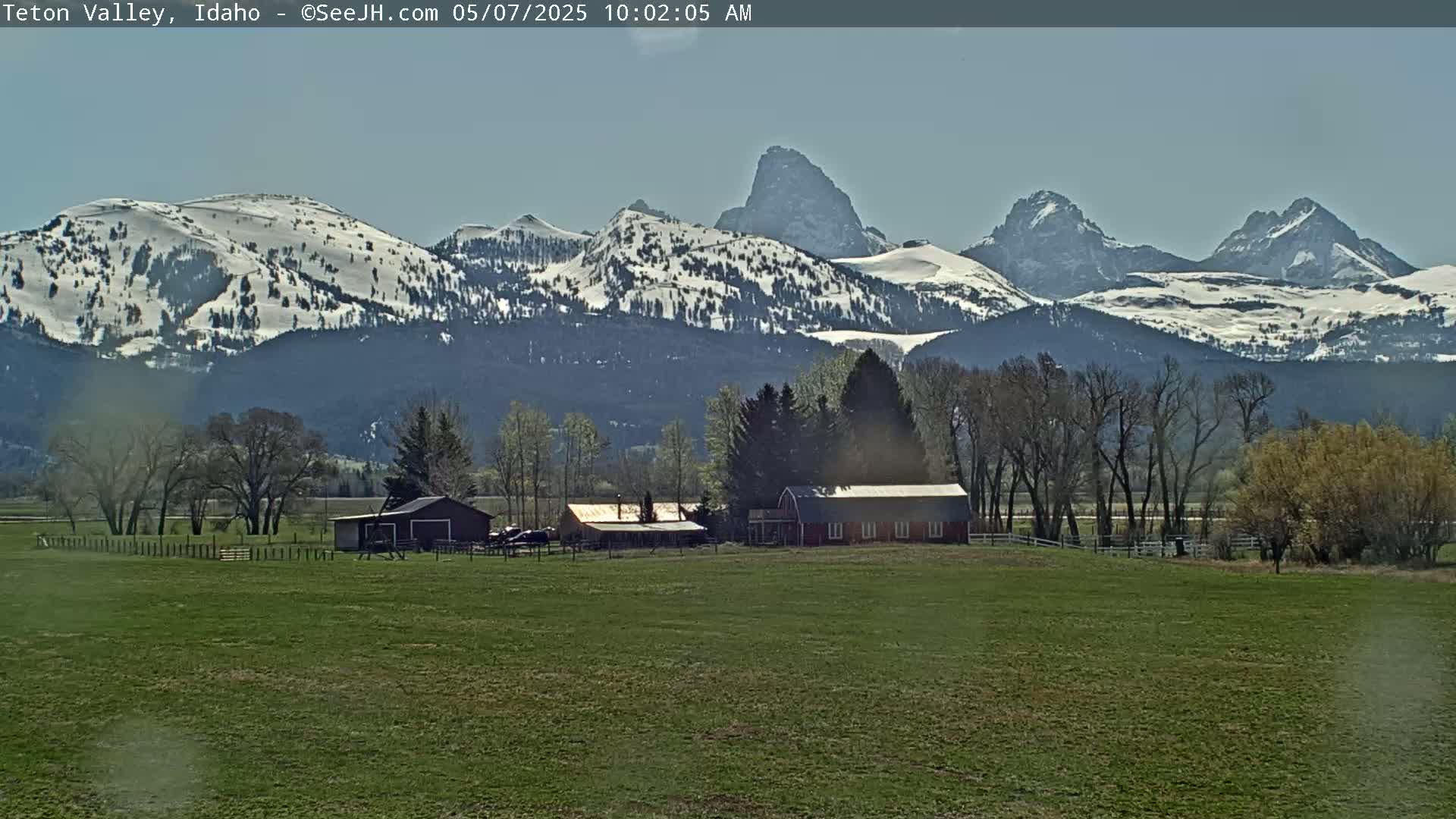 A red barn and other farm buildings sit on a grassy field, with snow-capped mountains in the background under a clear sky.