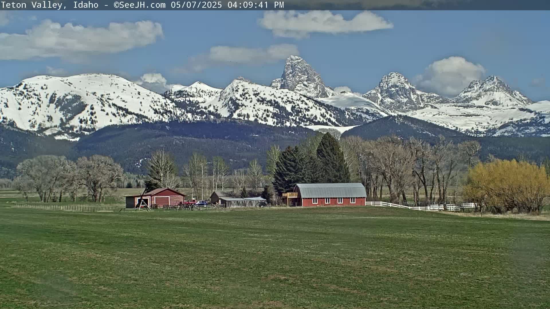 A verdant field stretches before a red barn and outbuildings, all set against a backdrop of snow-capped mountains under a partly cloudy blue sky.