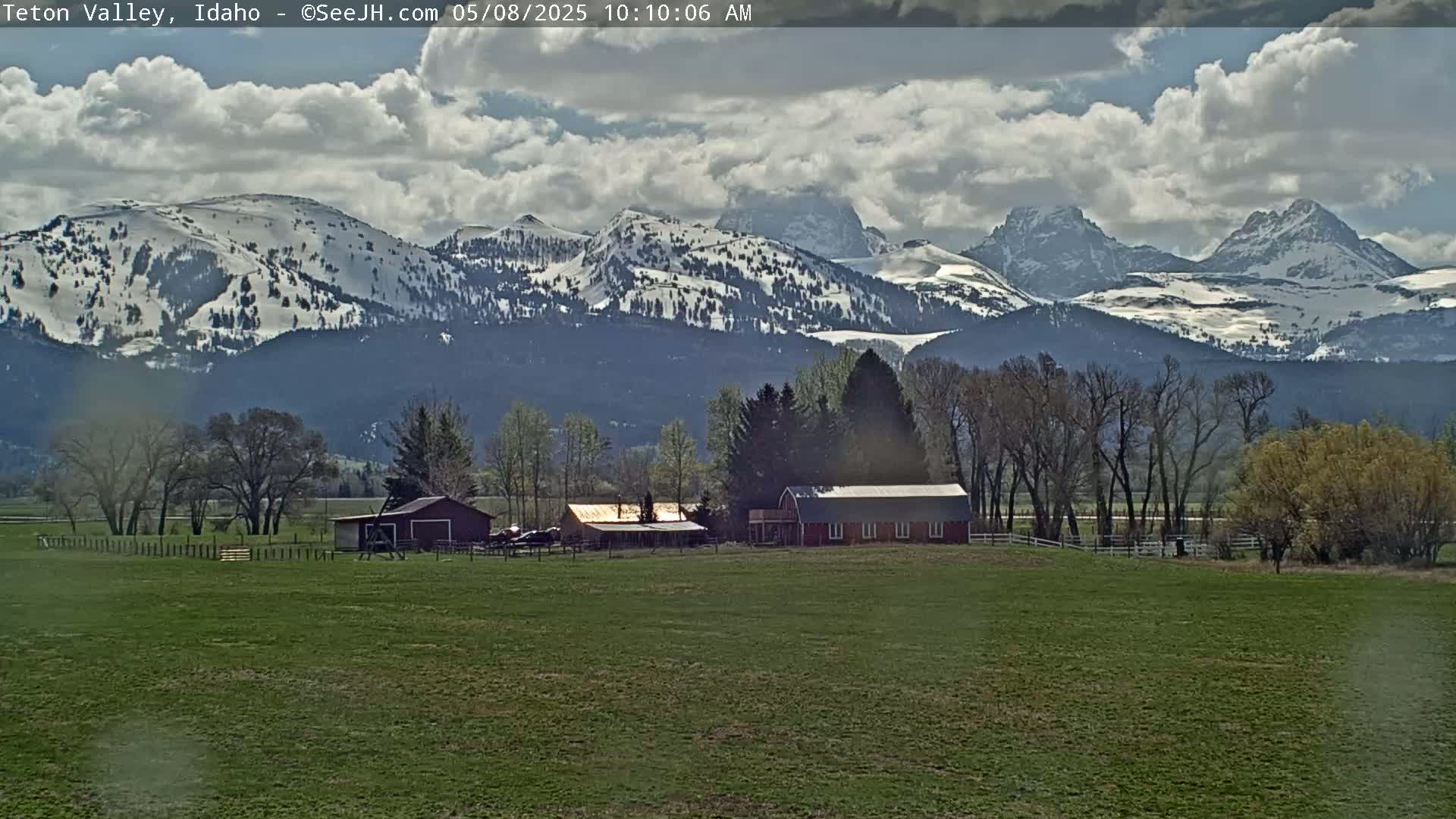 A partially snow-capped mountain range is visible in the background behind a farm with several buildings, under a partly cloudy sky.
