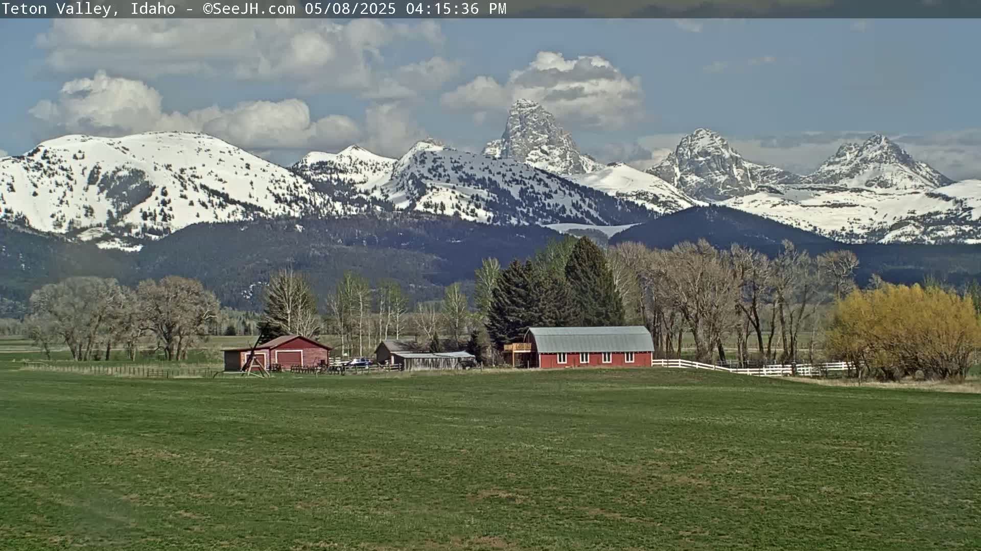A partly cloudy day shows snow-capped mountains in the background, with a green field in the foreground containing a red barn and other farm buildings.