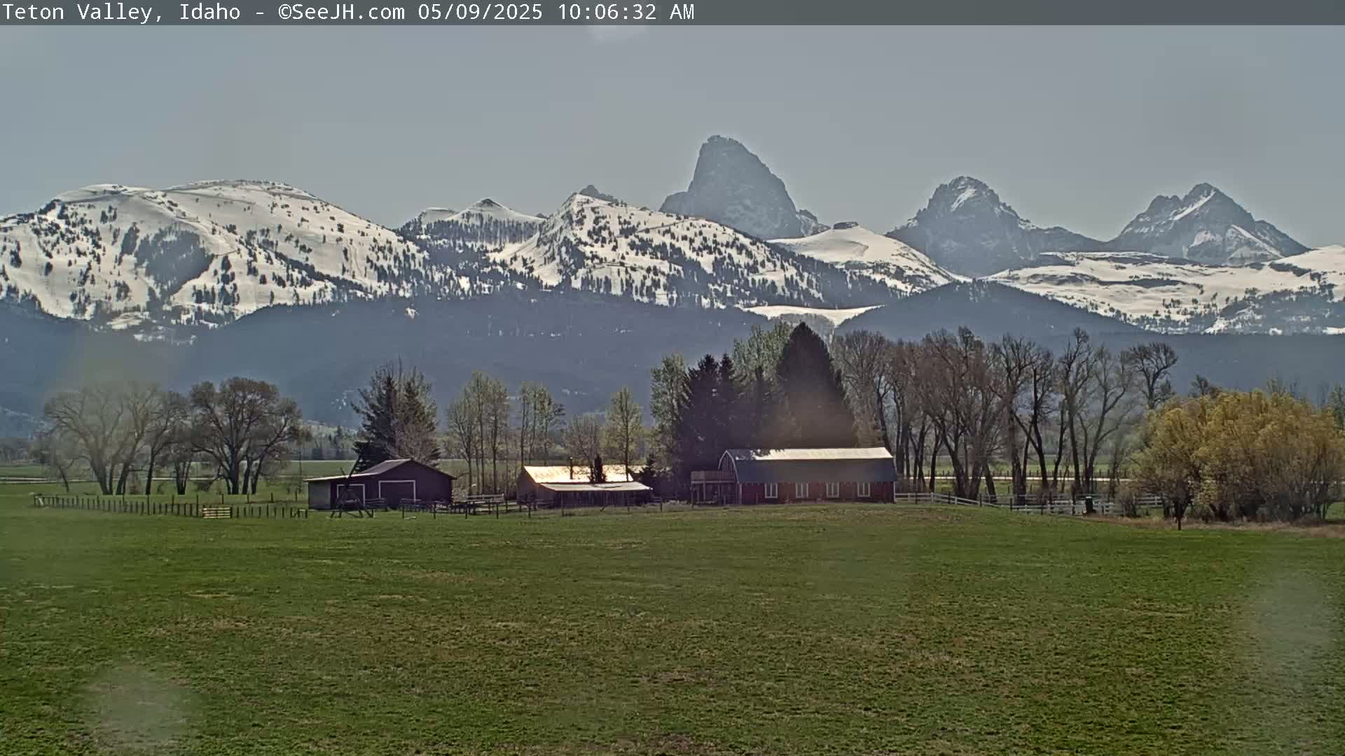 A farm with several buildings sits in a grassy field, with snow-capped mountains in the background under a clear sky.
