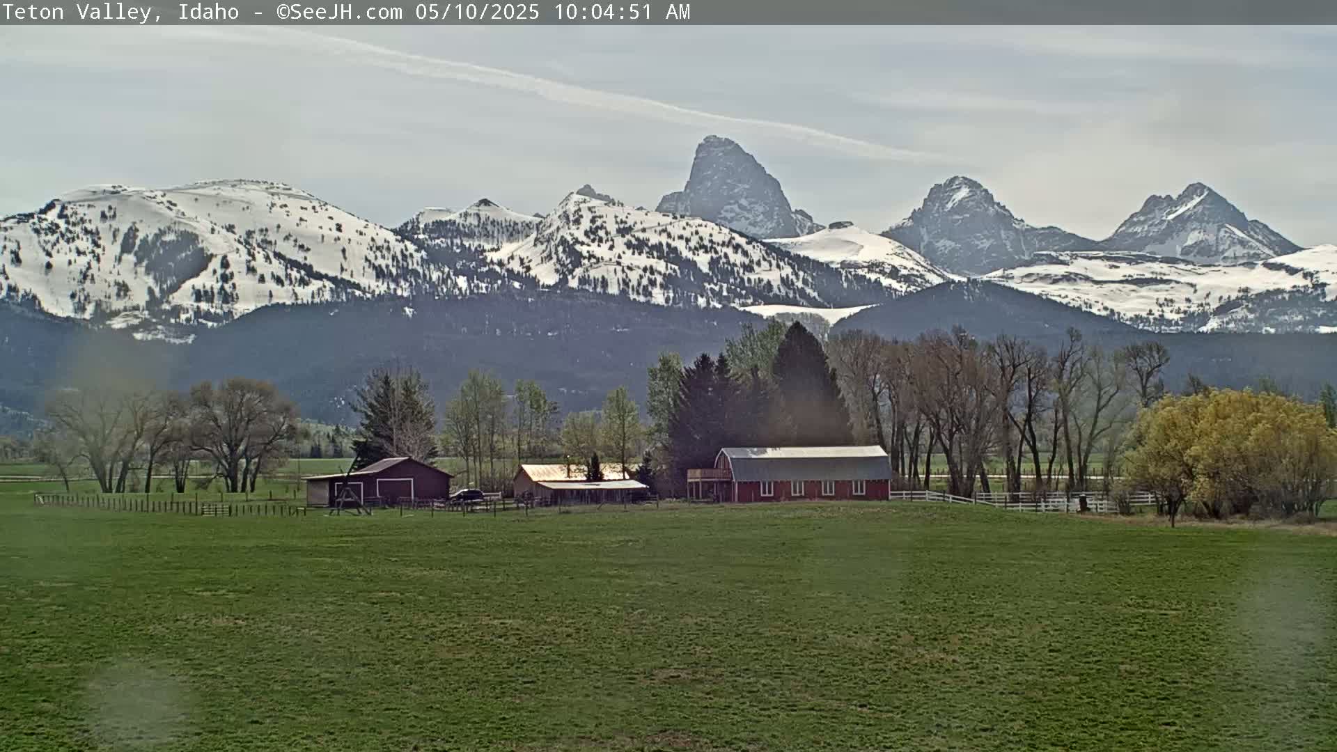 A red barn and other farm buildings sit in a green field, with snow-capped mountains in the background under a partly cloudy sky.