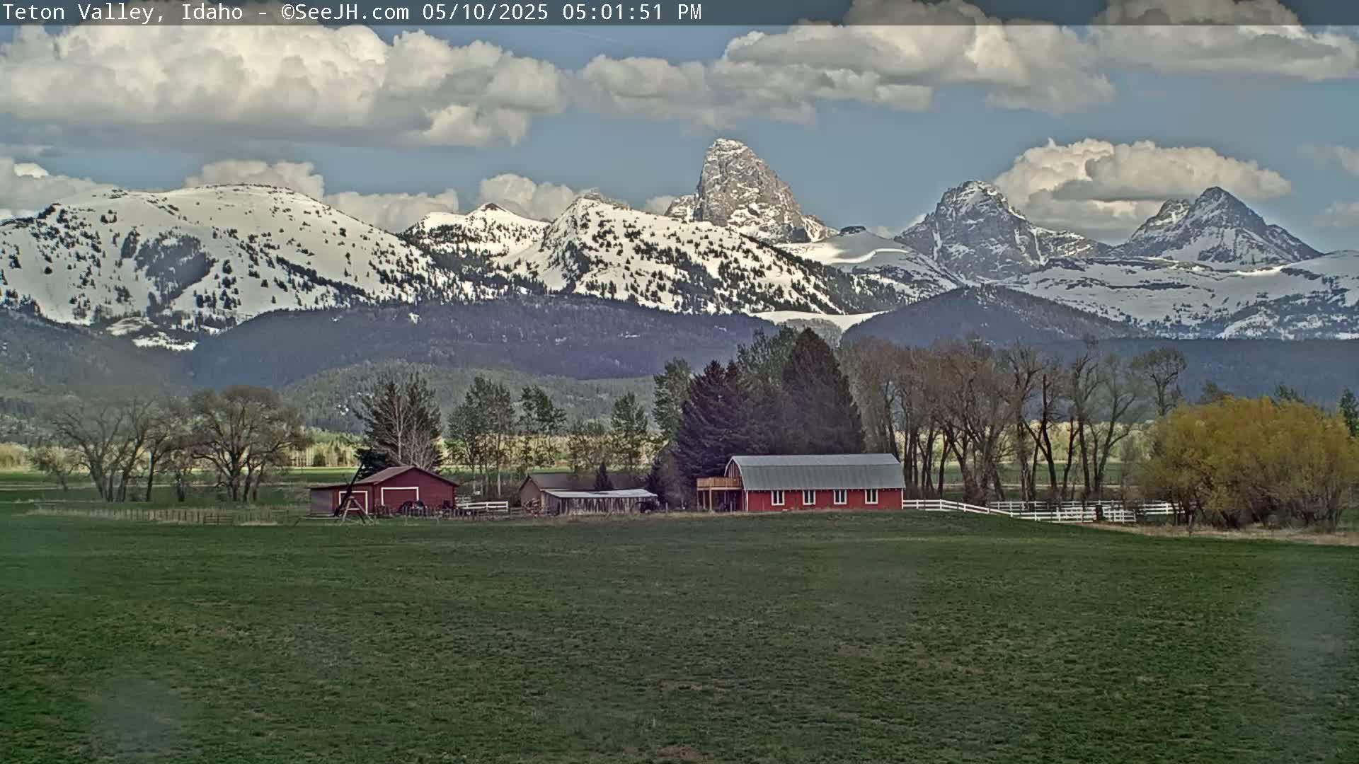 A red barn sits on a green field, with snow-capped mountains in the background under a partly cloudy sky.
