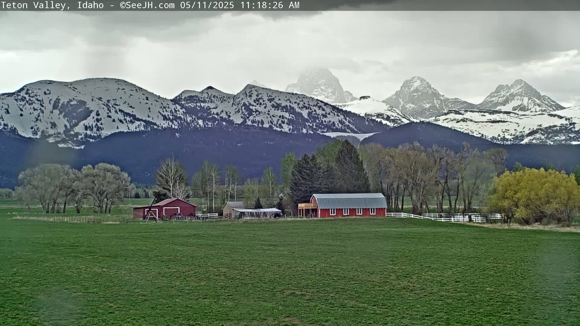 A red barn sits on a green field in front of snow-capped mountains under an overcast sky.