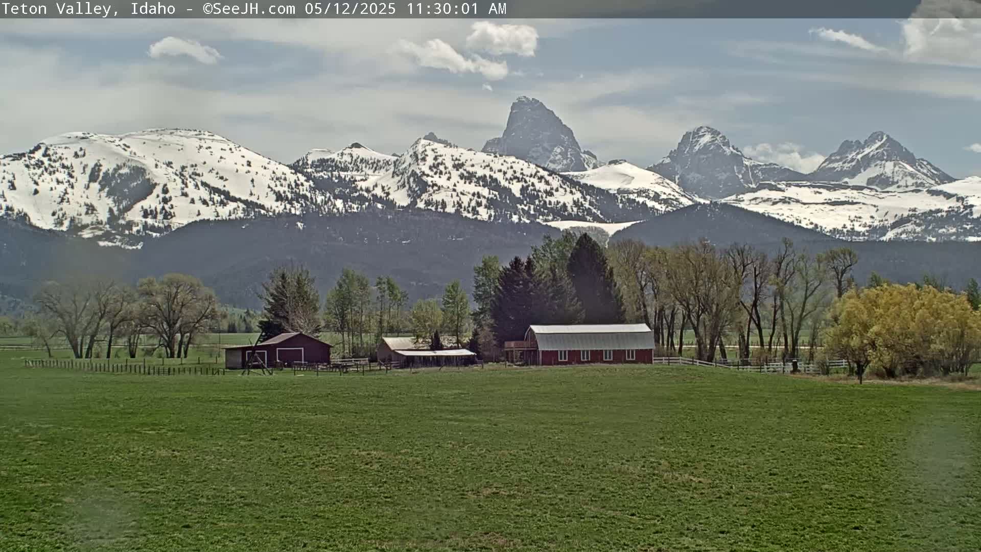 A partly cloudy day reveals snow-capped mountains in the distance behind a green field with a red barn and other farm buildings.