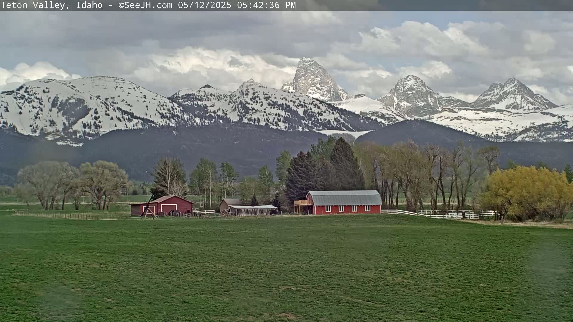 A partly cloudy day reveals a snow-capped mountain range behind a green field, where a red barn and other farm buildings sit among leafed trees.