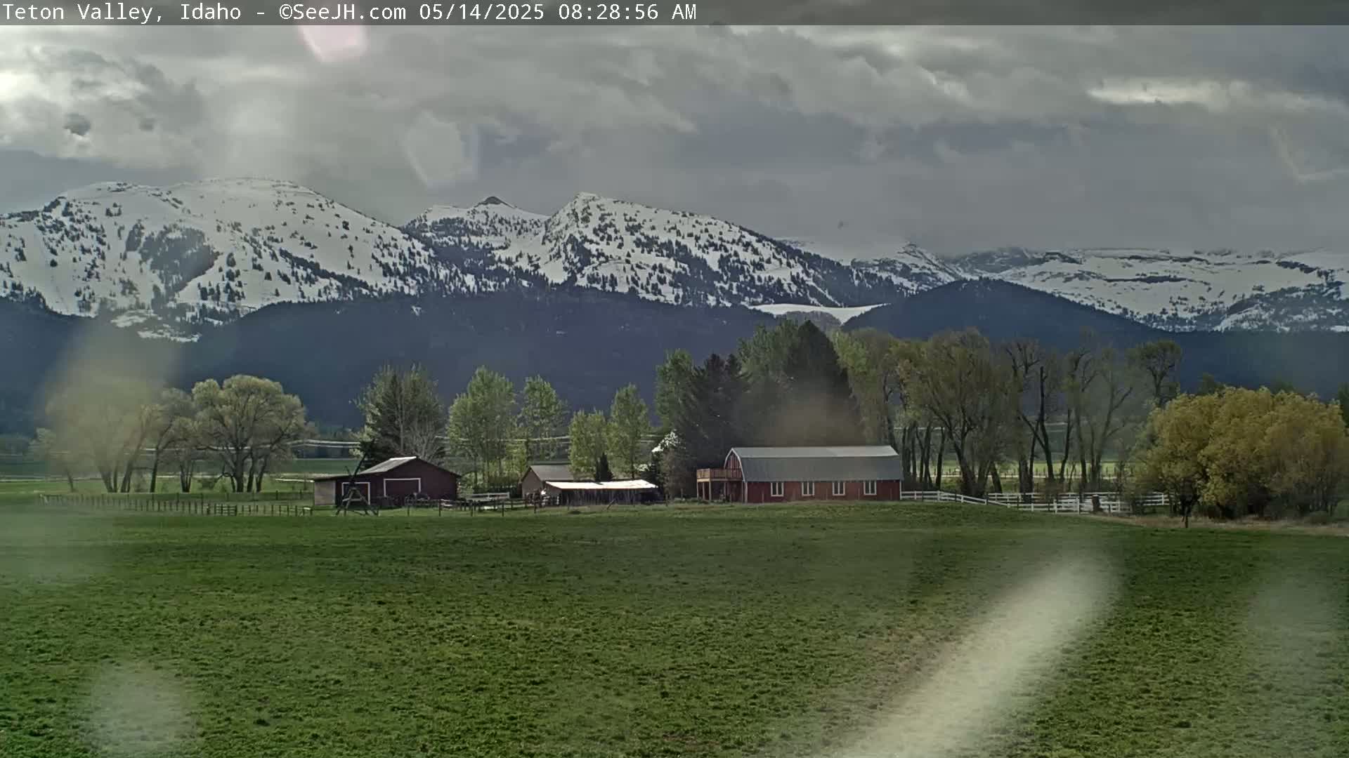 A green pasture with a red barn and other farm buildings sits in front of snow-capped mountains under a cloudy sky.