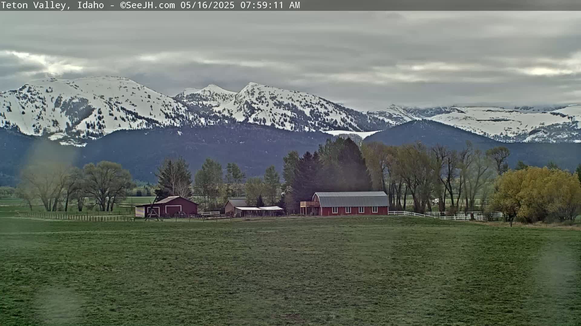 A red barn and other farm buildings sit in a green field, with snow-capped mountains in the background under a cloudy sky.