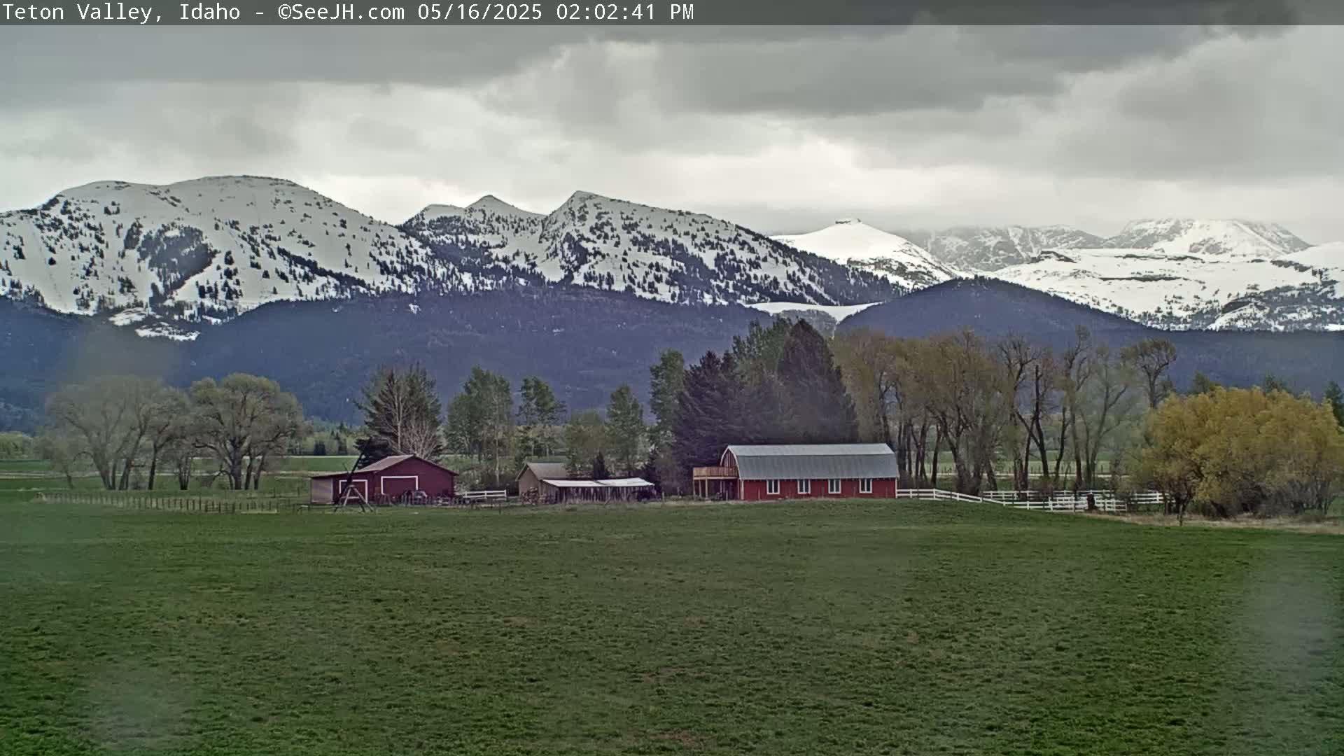 A green field with a red barn and other outbuildings sits before snow-capped mountains under a cloudy sky.