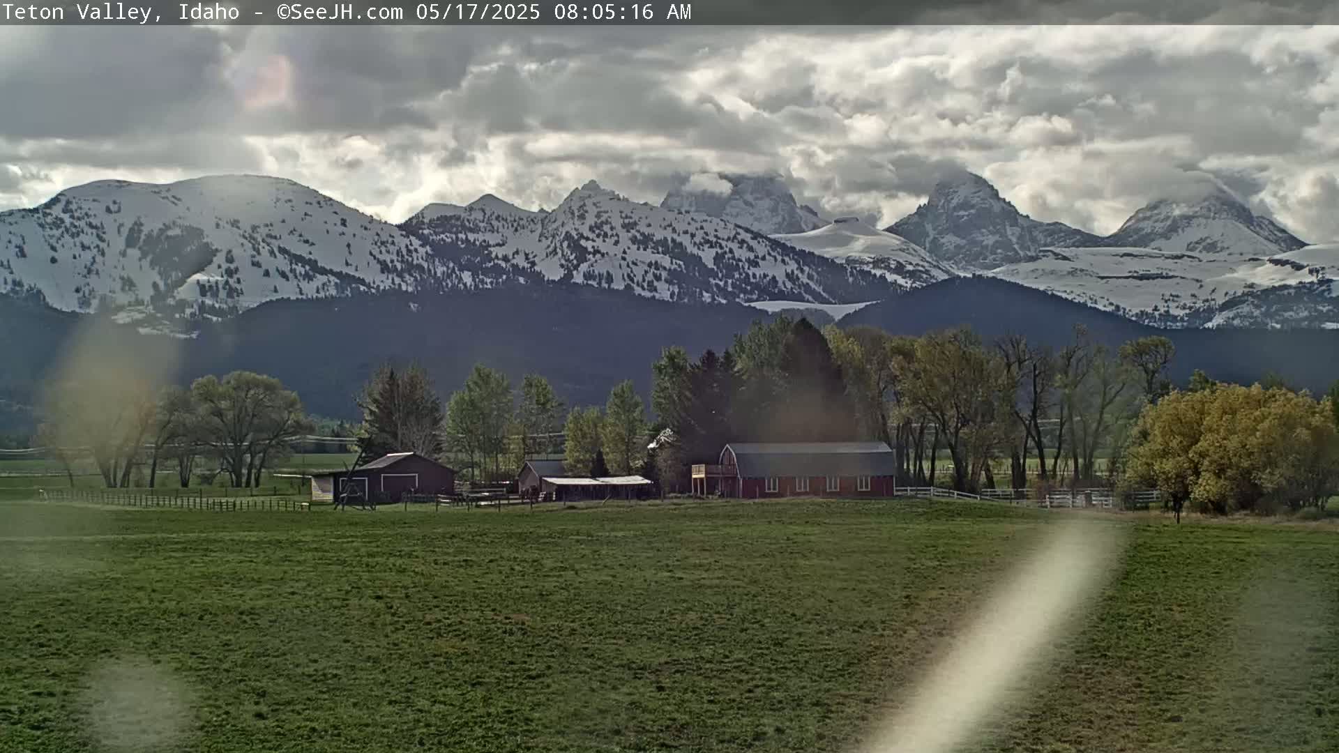 A farm with several buildings sits in a verdant field, backed by snow-capped mountains under a partly cloudy sky.