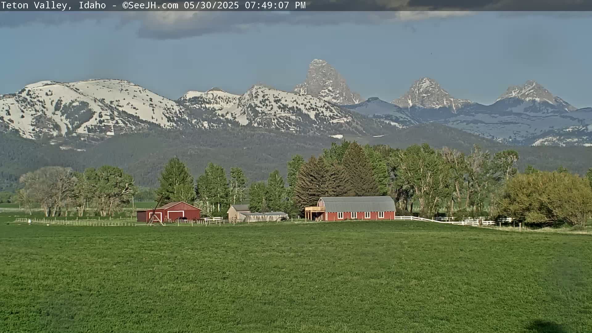 A red barn sits on a green field with a line of trees in front of snow-capped mountains under a mostly clear sky.