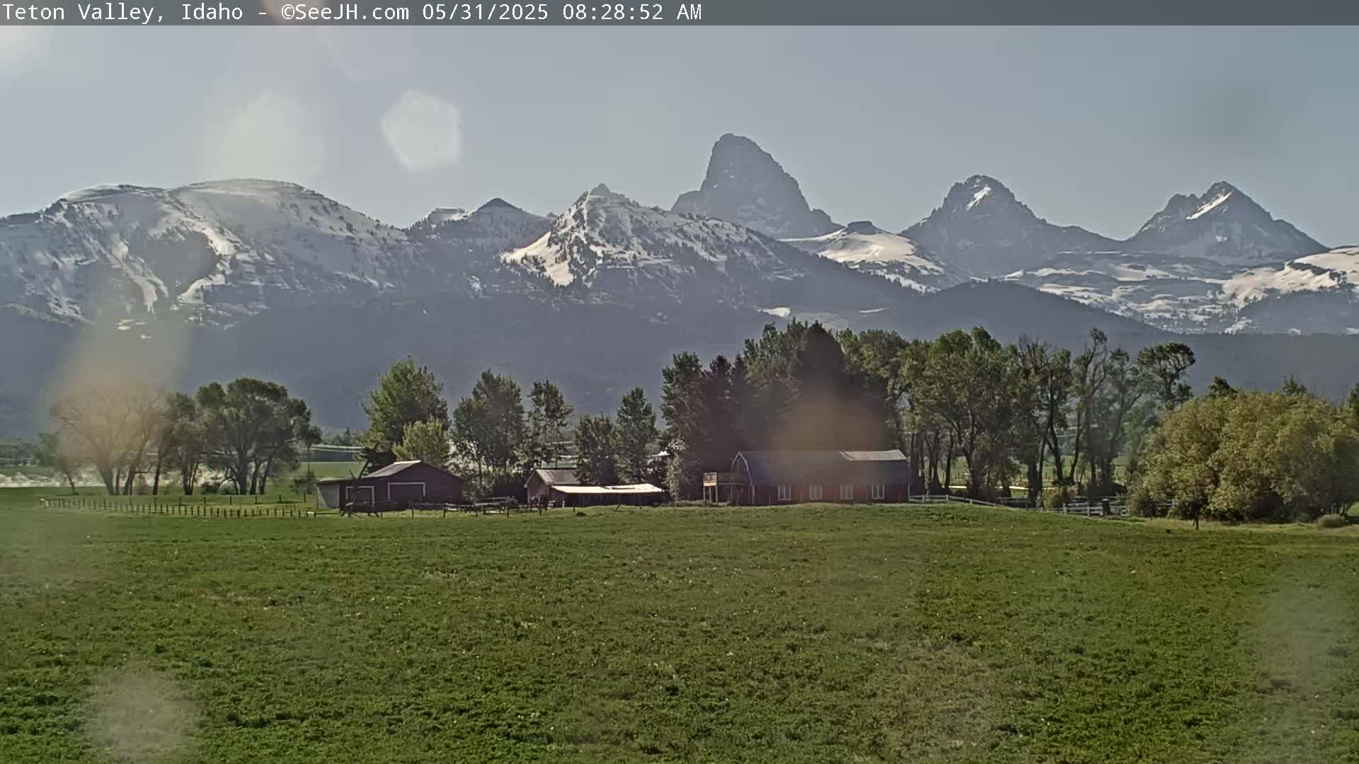 A green field with farm buildings sits in the foreground of a snow-capped mountain range under a clear sky.