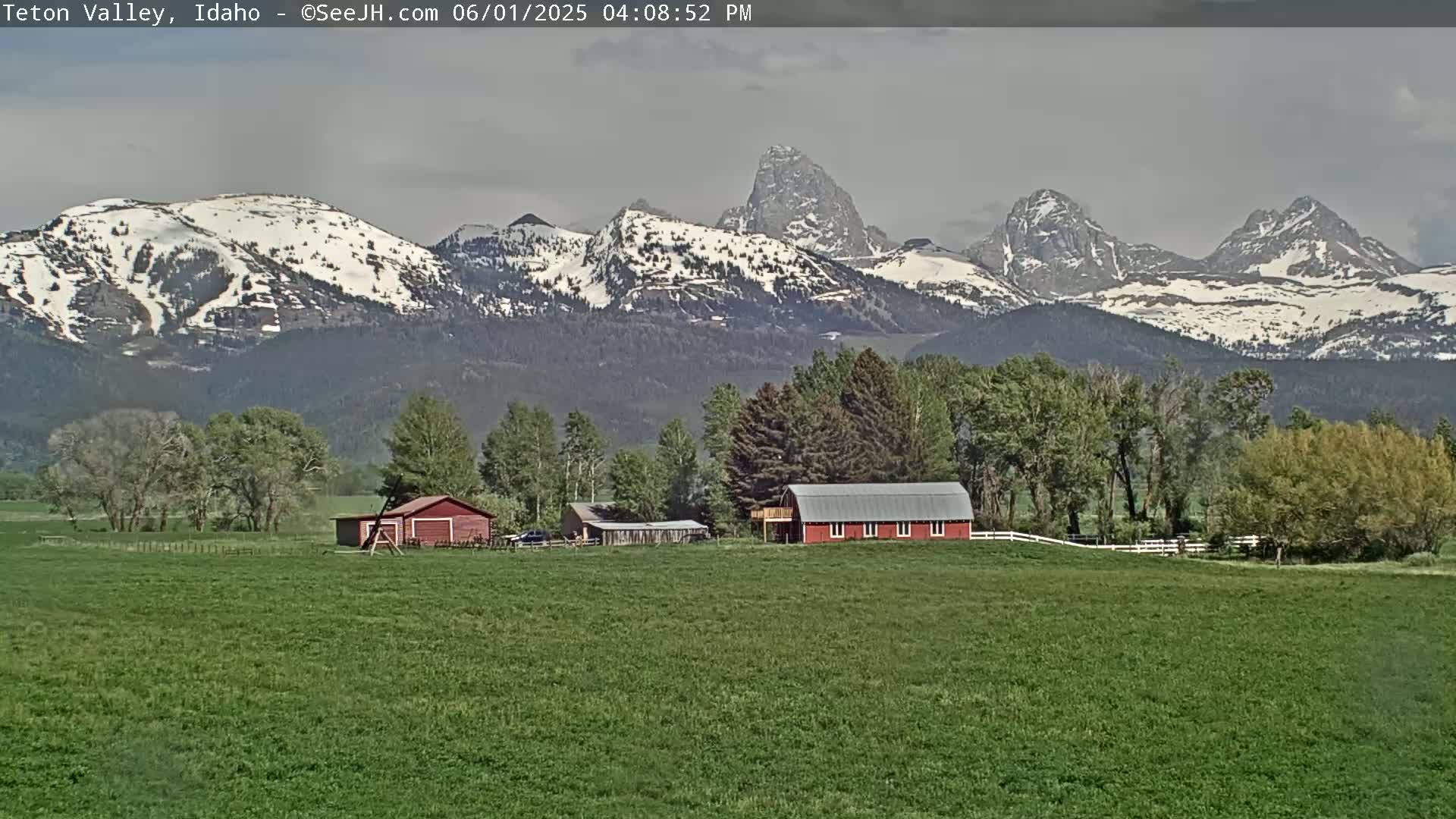 A red barn sits in a green field, with snow-capped mountains in the background under a mostly cloudy sky.