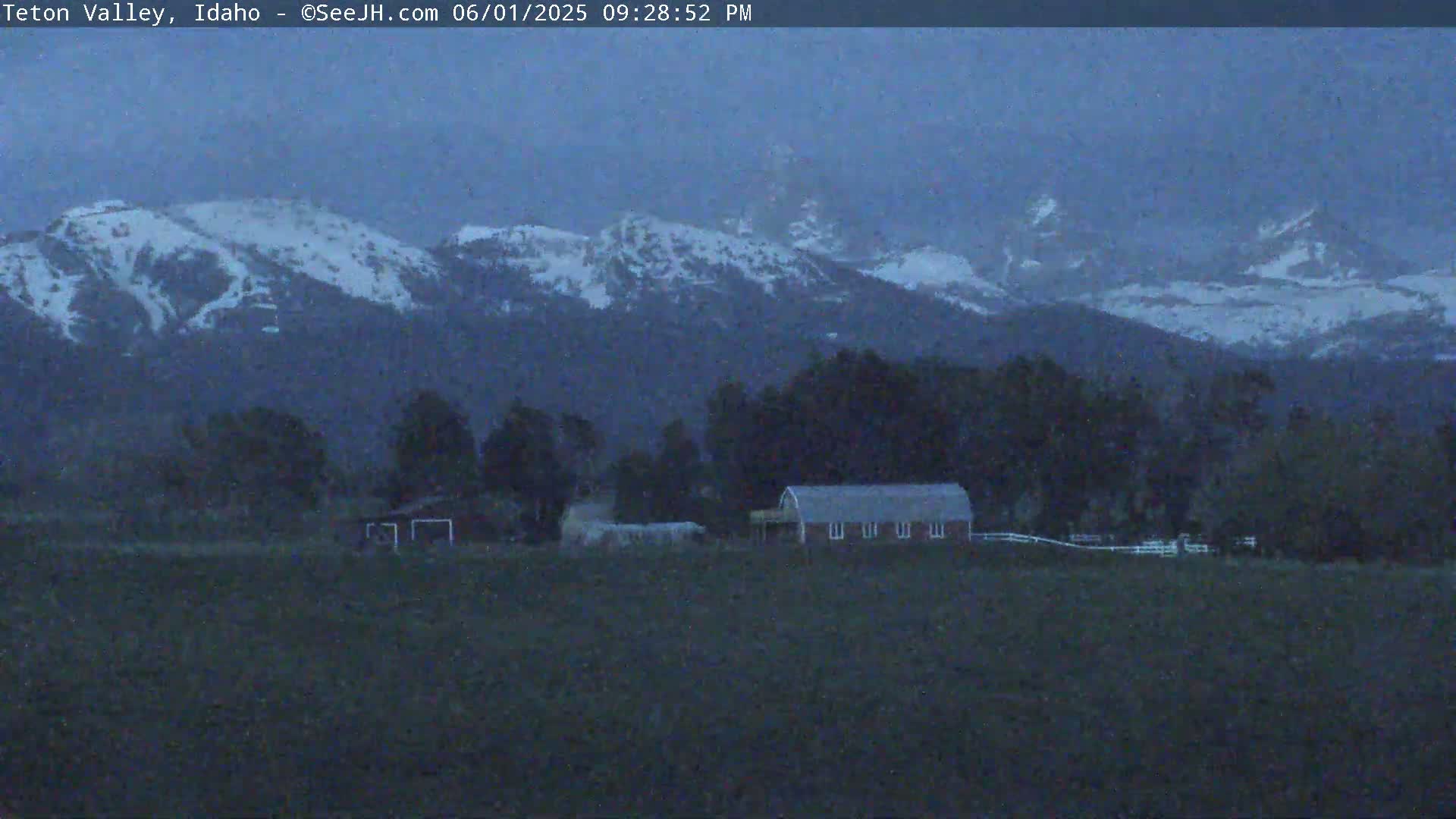 Snow-capped mountains loom in the dusky twilight over a flat expanse of land containing a red barn and other structures.