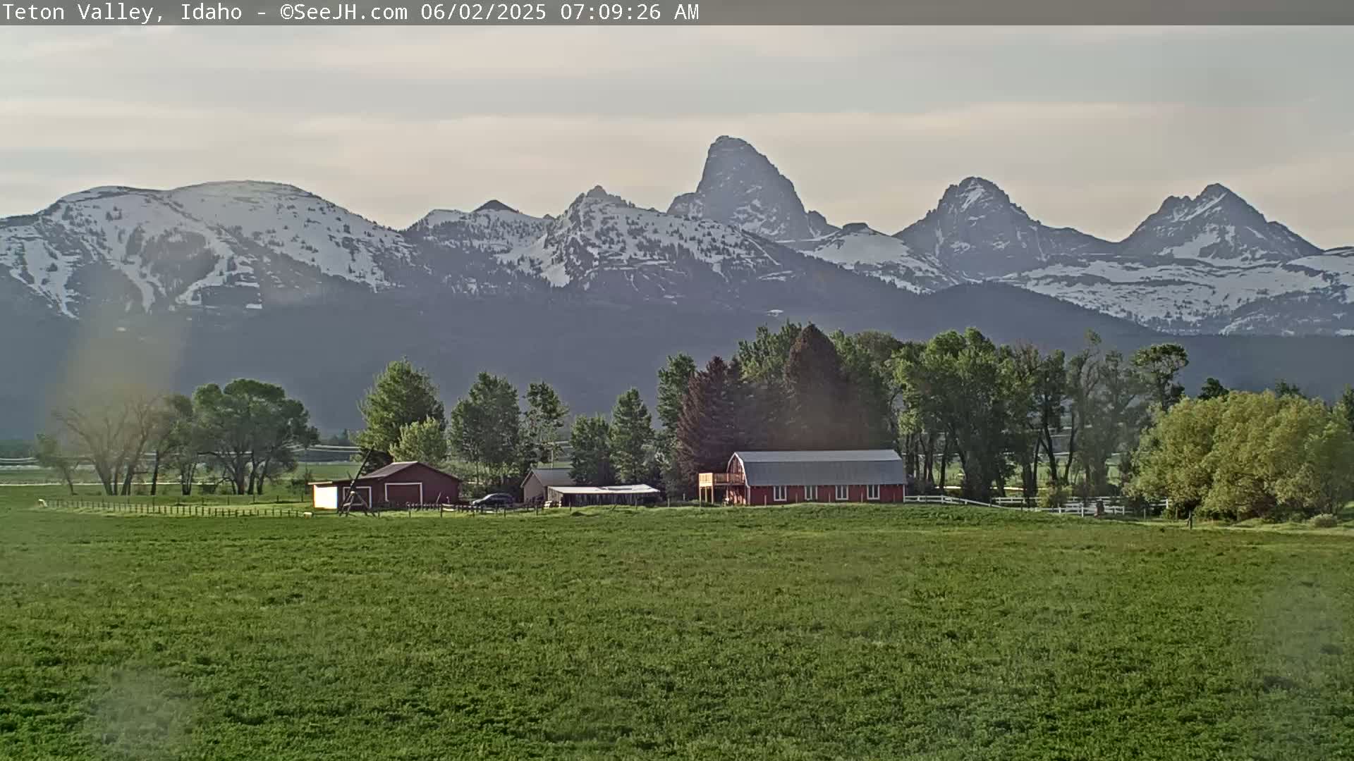 A red barn and other farm buildings sit on a grassy field in front of a snow-capped mountain range under a mostly cloudy sky.