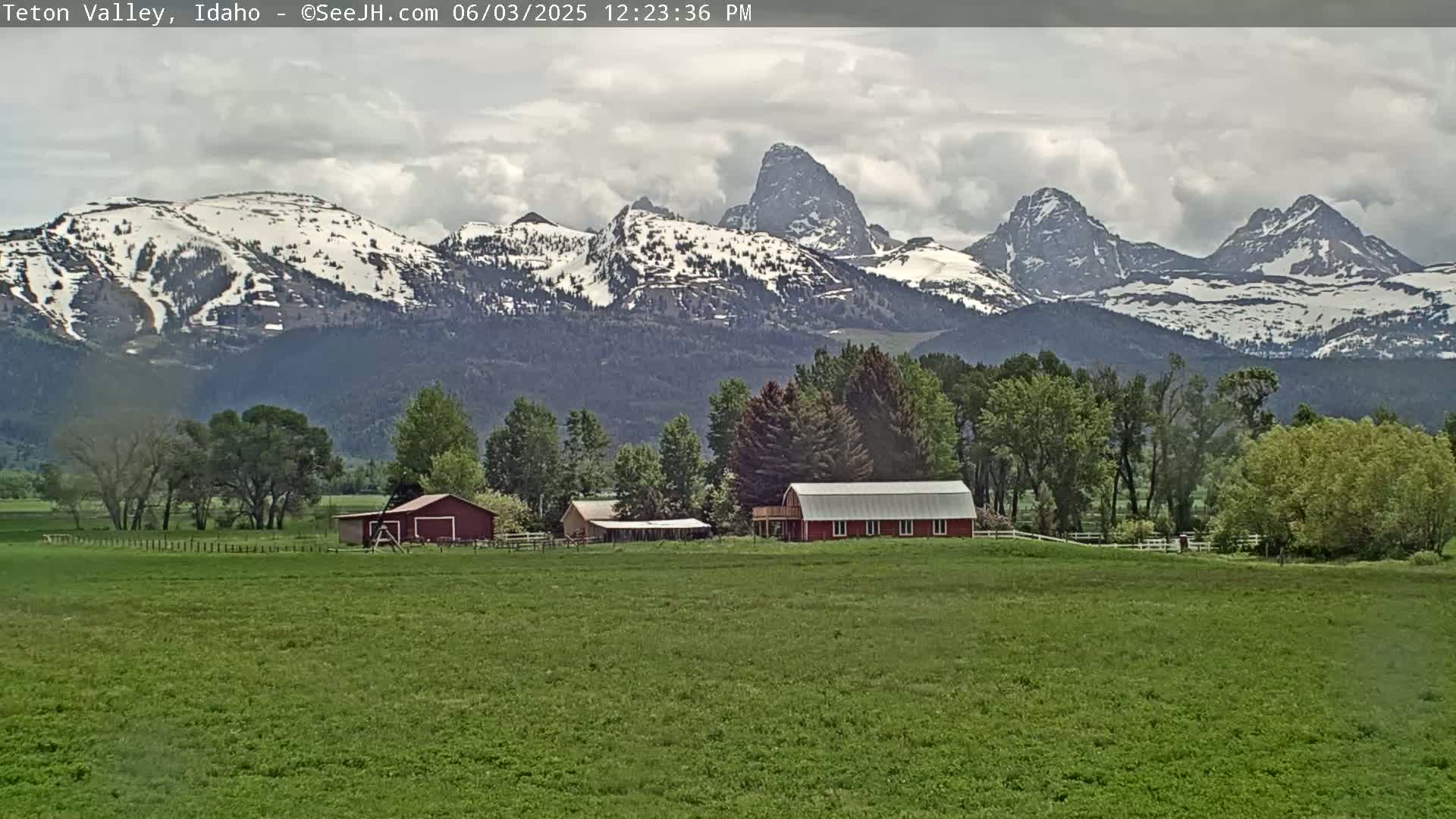 A green field with red barns sits in front of snow-capped mountains under a cloudy sky.