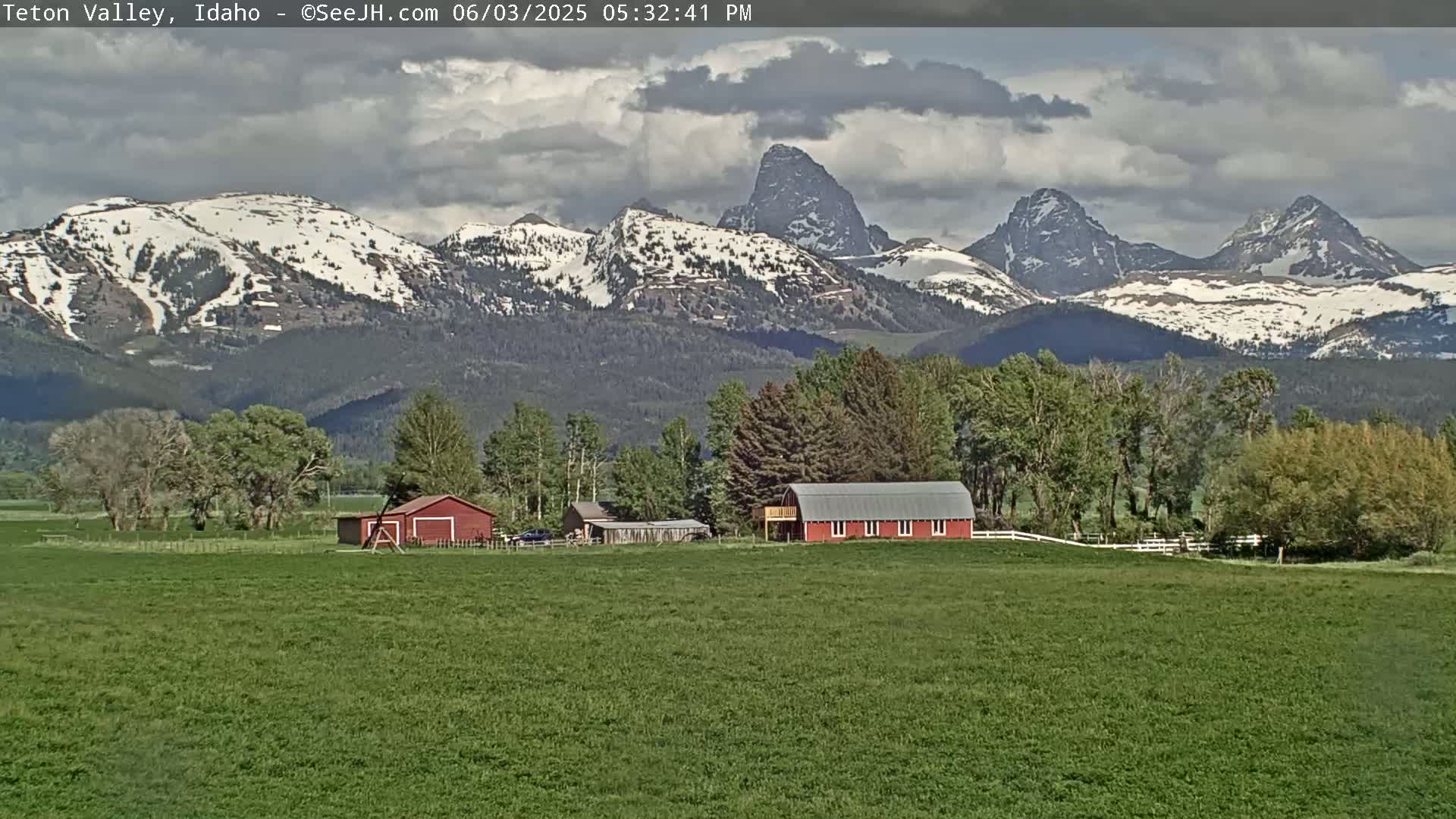 A green field with two red barns sits in front of a mountain range with snow-capped peaks under a partly cloudy sky.