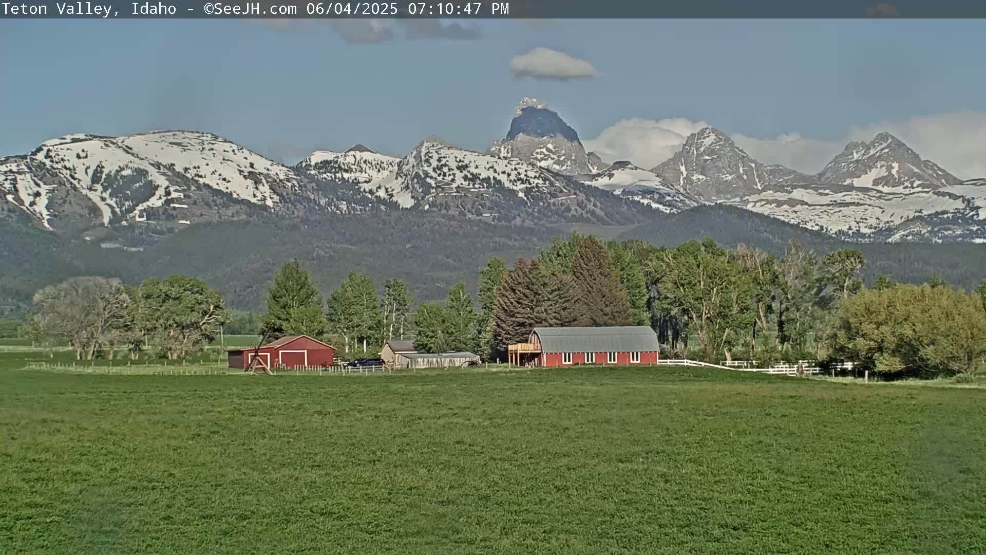 A green field with a red barn and other buildings sits in front of snow-capped mountains under a partly cloudy sky.