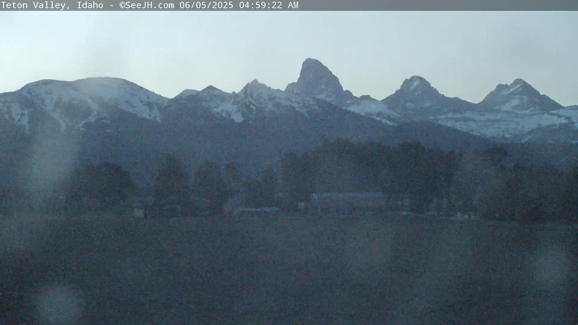 A hazy, pre-dawn view shows a snow-capped mountain range in the distance, with a flat, dark foreground containing some low-lying vegetation.