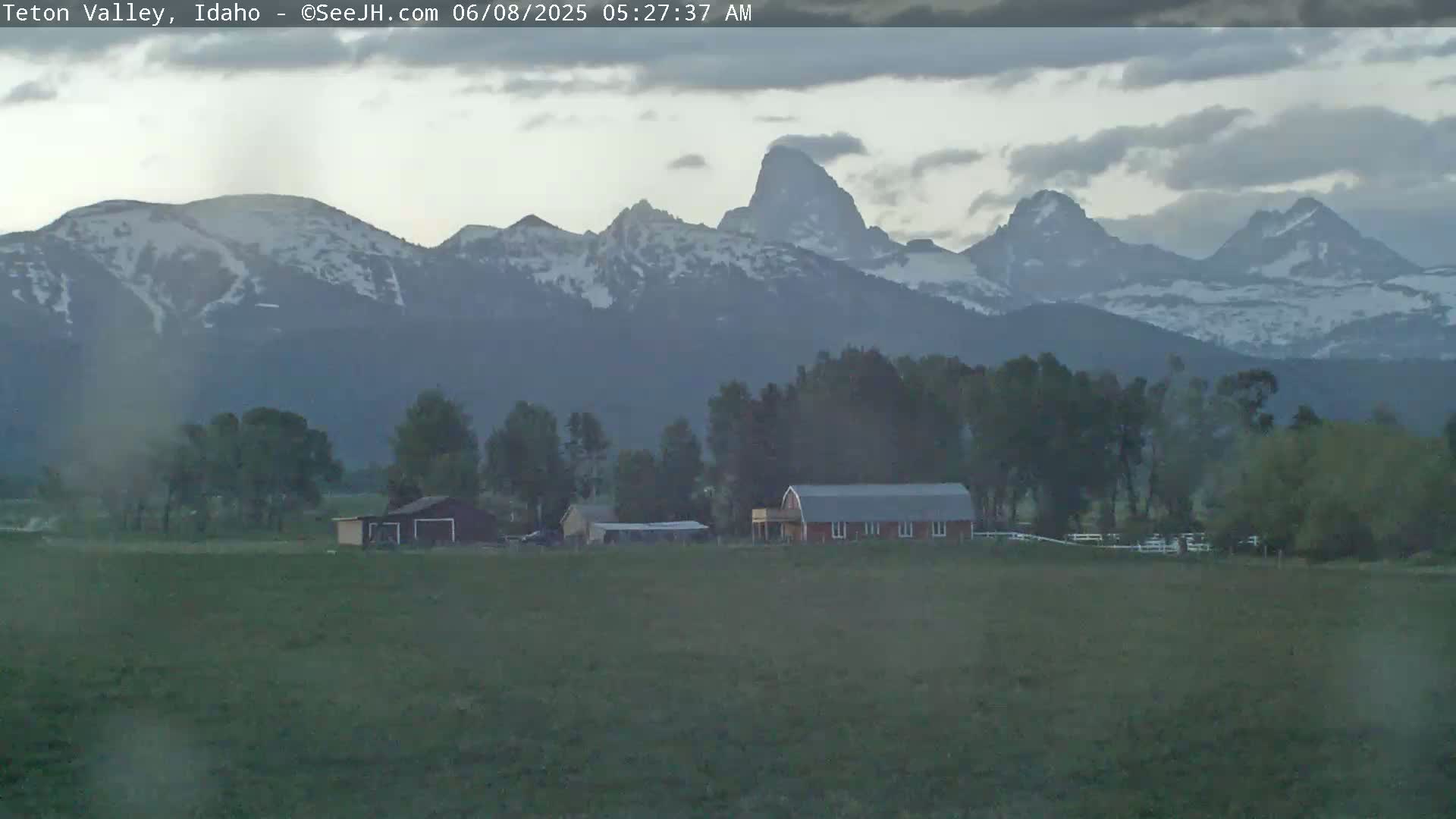A farm with a red barn sits in a field, with snow-capped mountains in the background under a cloudy sky.