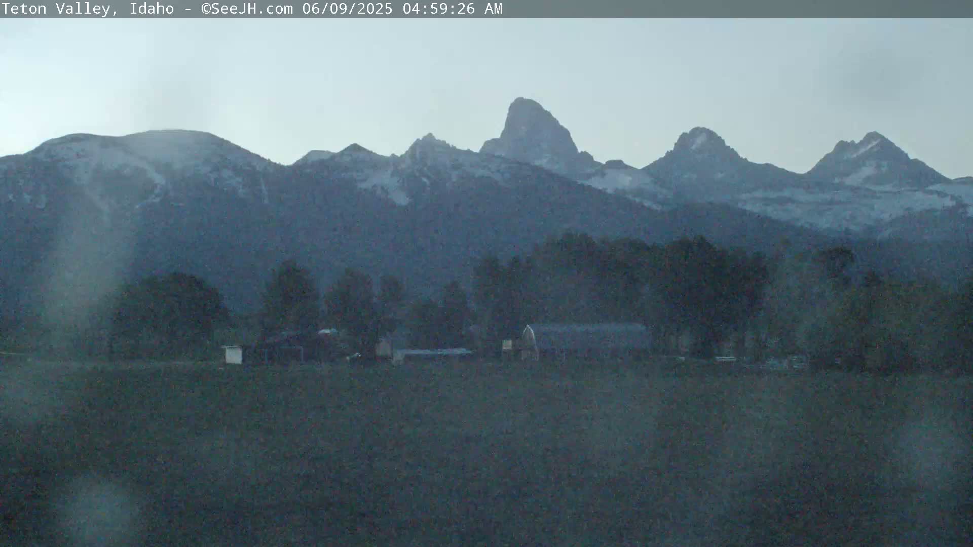 A hazy, pre-dawn view shows snow-capped mountains behind a flat field with a few buildings and trees.