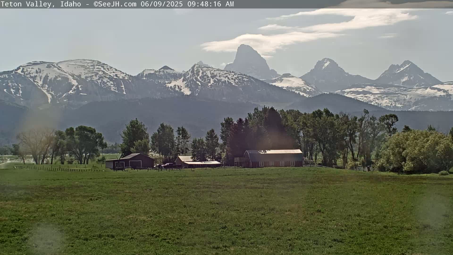 A partly cloudy day reveals a grassy field with farm buildings in the foreground and snow-capped mountains in the background.