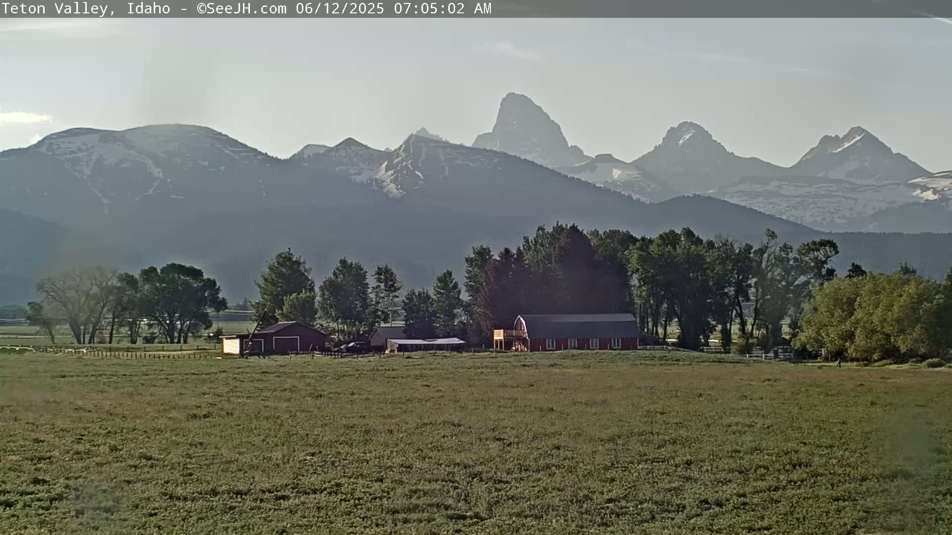 A green field with a red barn and other buildings sits in the foreground, with snow-capped mountains rising in the background under a hazy sky.