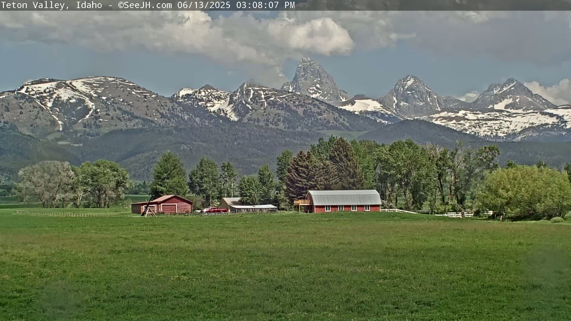 A partly cloudy day reveals a green field with a red barn and other farm buildings in the foreground, set against a backdrop of snow-capped mountains.