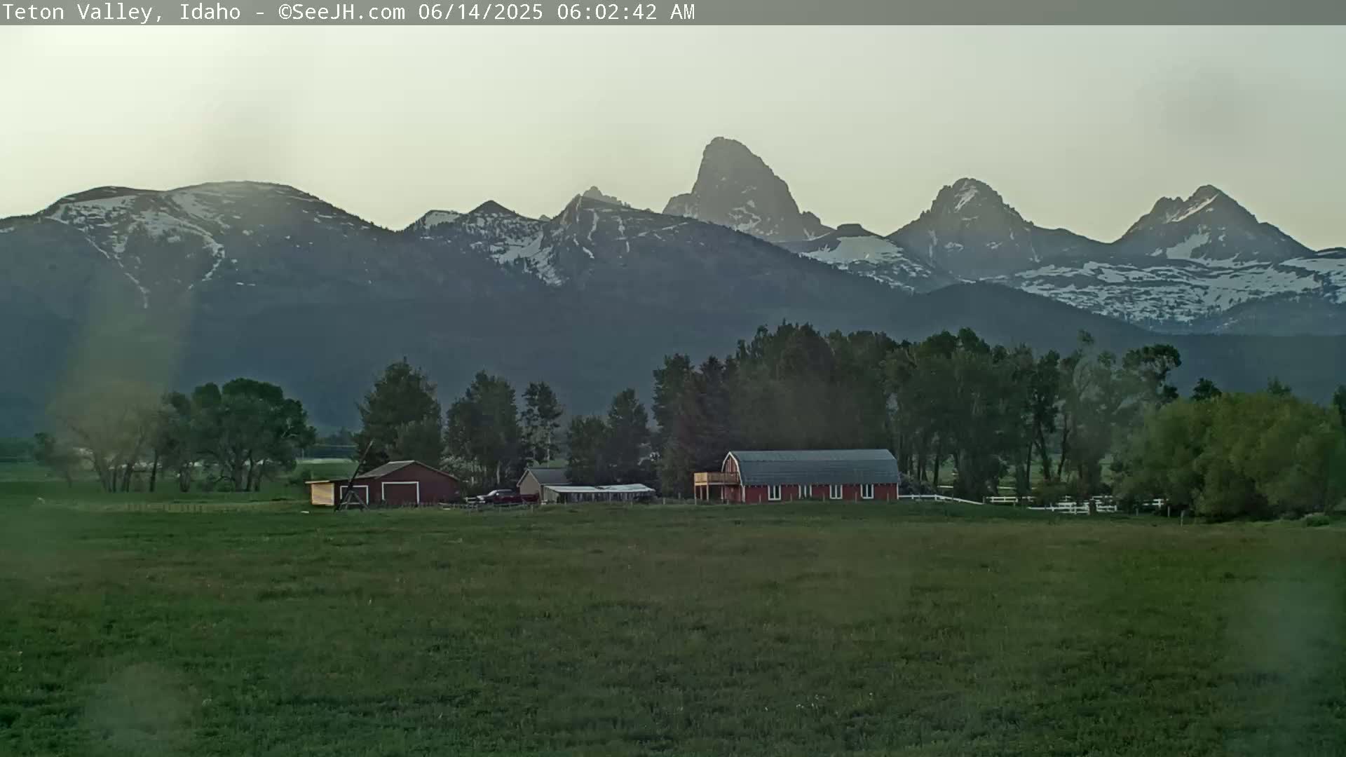 A red barn and other farm buildings sit on a green field in front of a snow-capped mountain range under a twilight sky.
