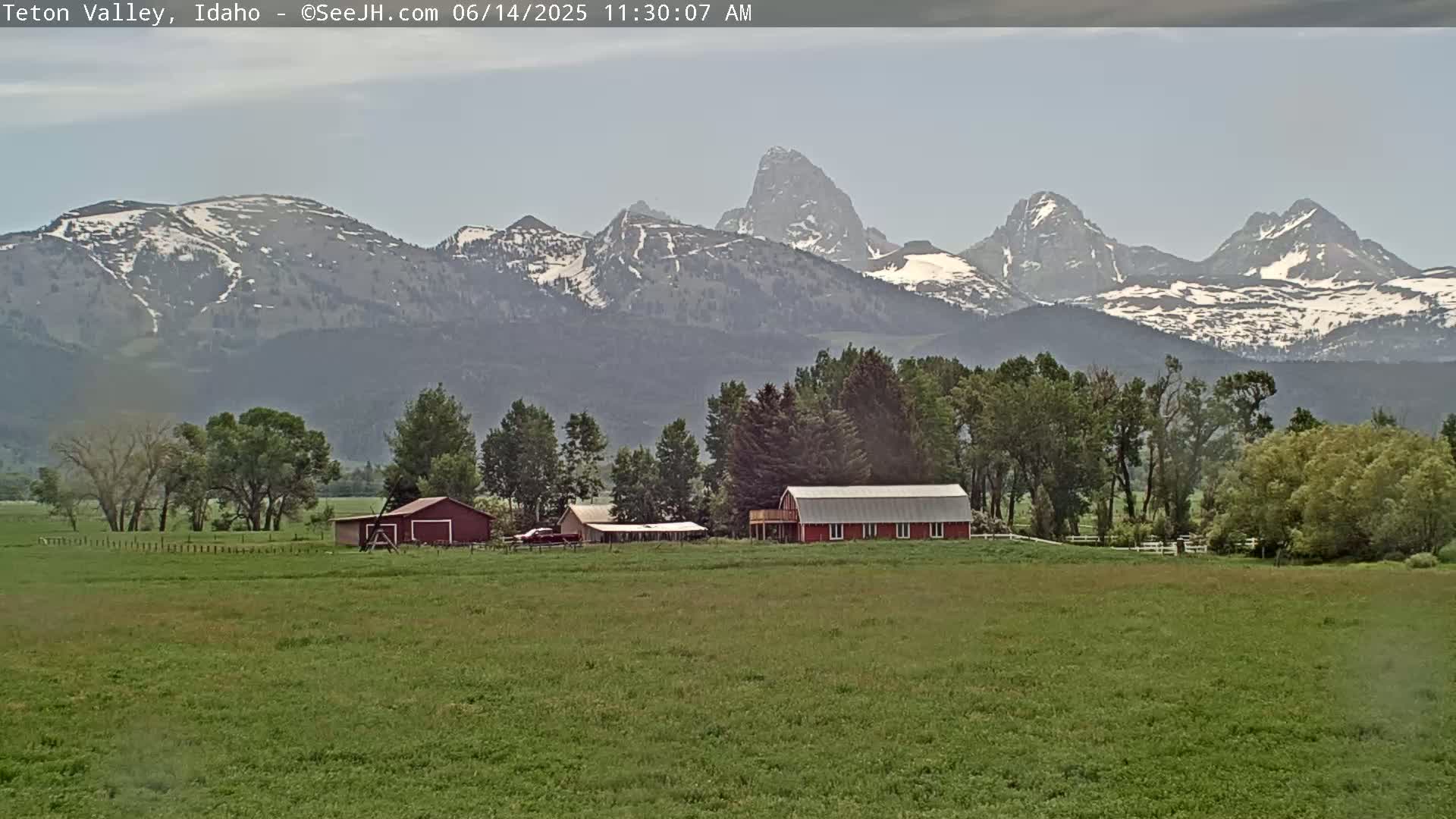 A red barn sits on a green field in front of a mountain range with patches of snow, under a mostly cloudy sky.