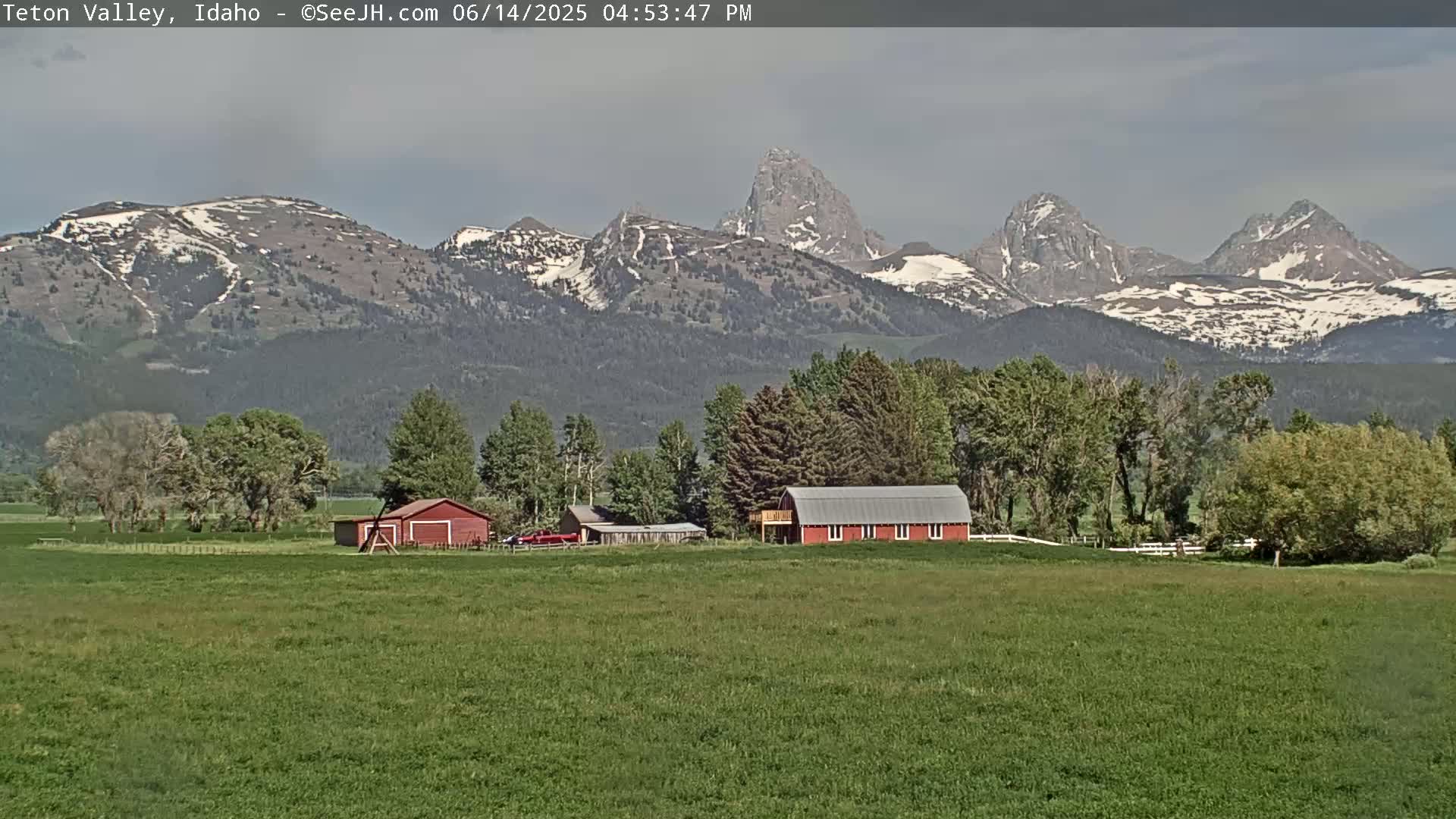A red barn sits on a green field in front of a mountain range with patches of snow, under a mostly cloudy sky.