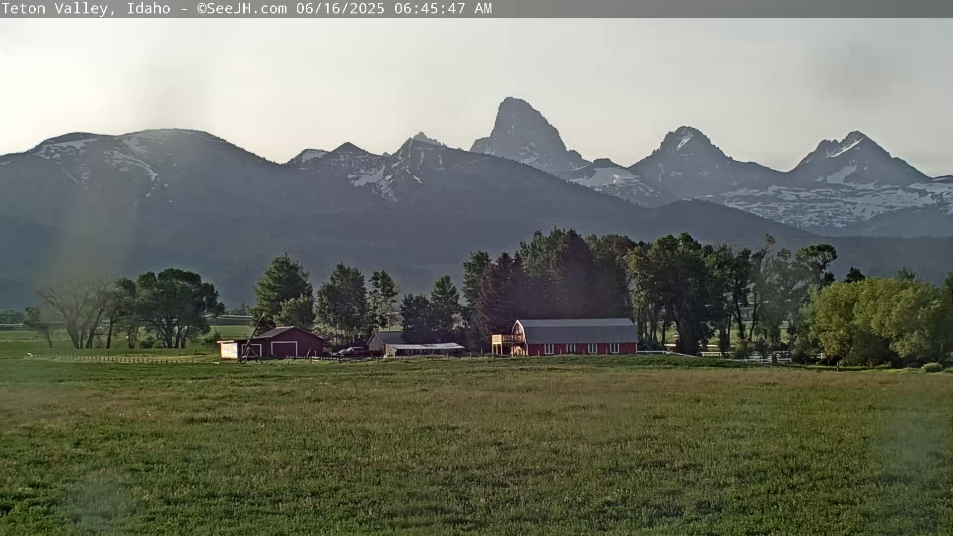 A red barn sits on a green field in front of a mountain range with some snow, under a mostly clear sky.