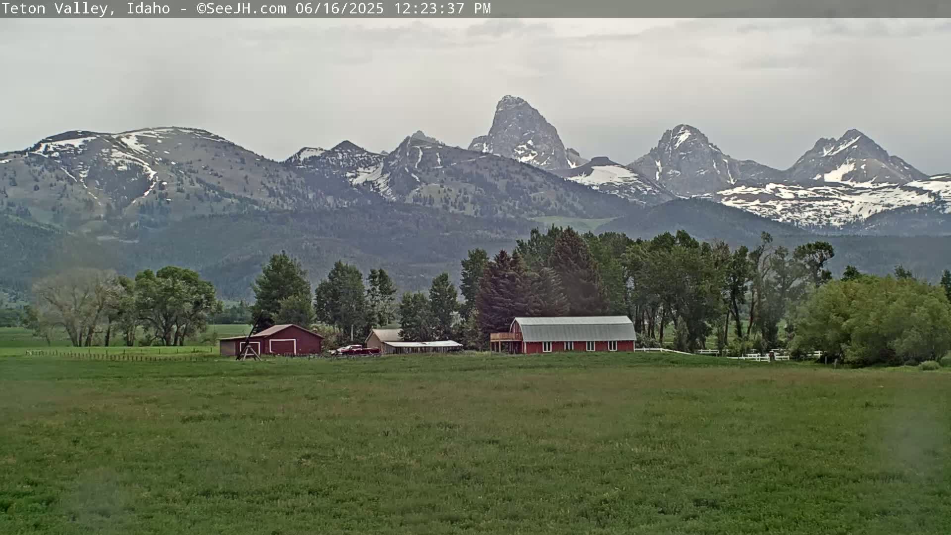 A red barn sits on a green field, with snow-capped mountains in the background under an overcast sky.