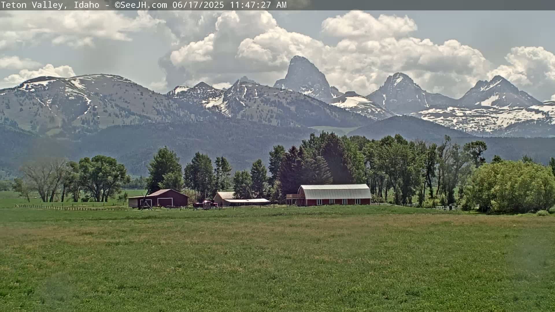 A partly cloudy day reveals a green field with farm buildings in the foreground and snow-capped mountains in the background.