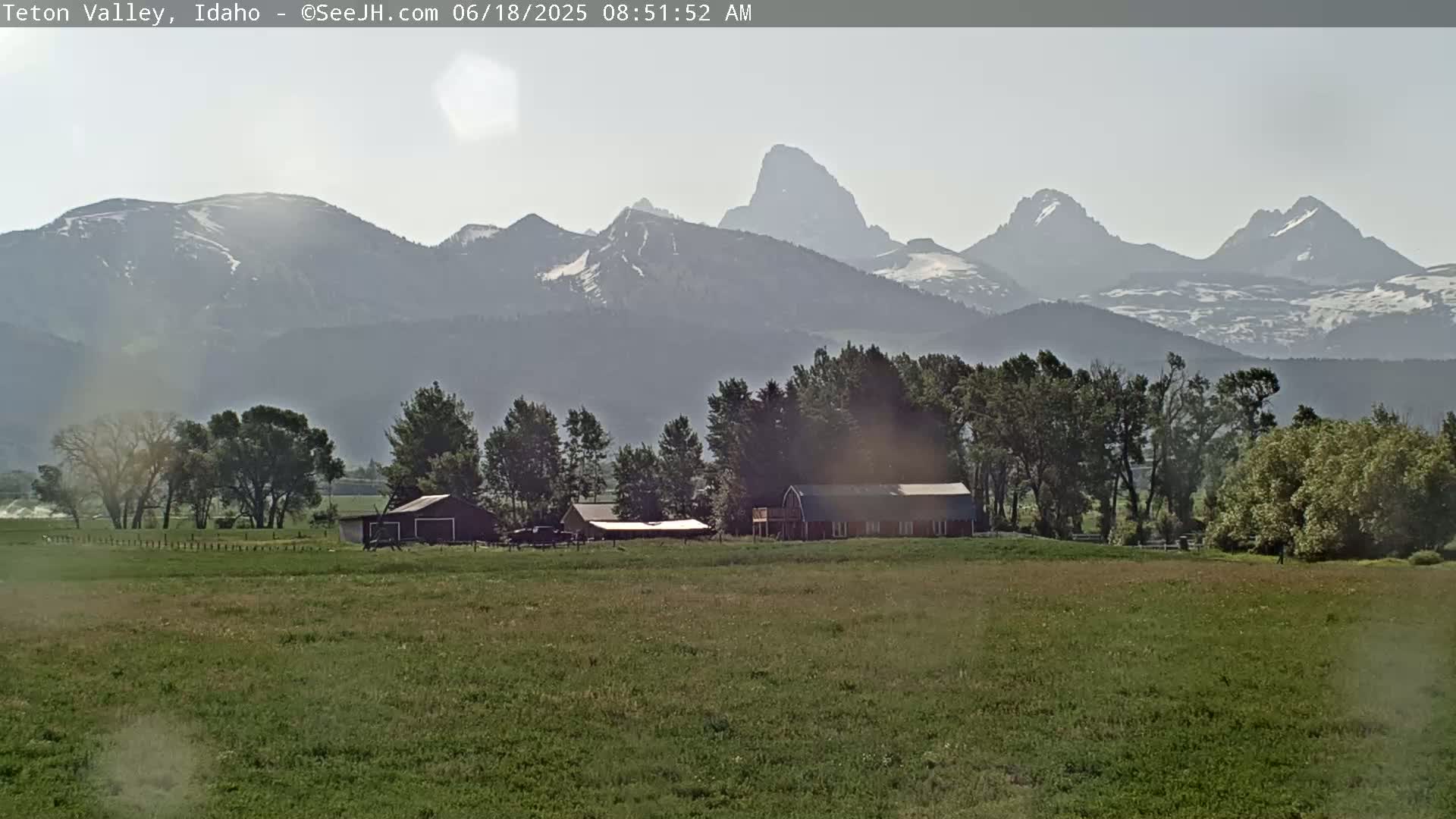 A grassy field with a few buildings sits in front of a hazy mountain range with some snow-capped peaks under a partly sunny sky.