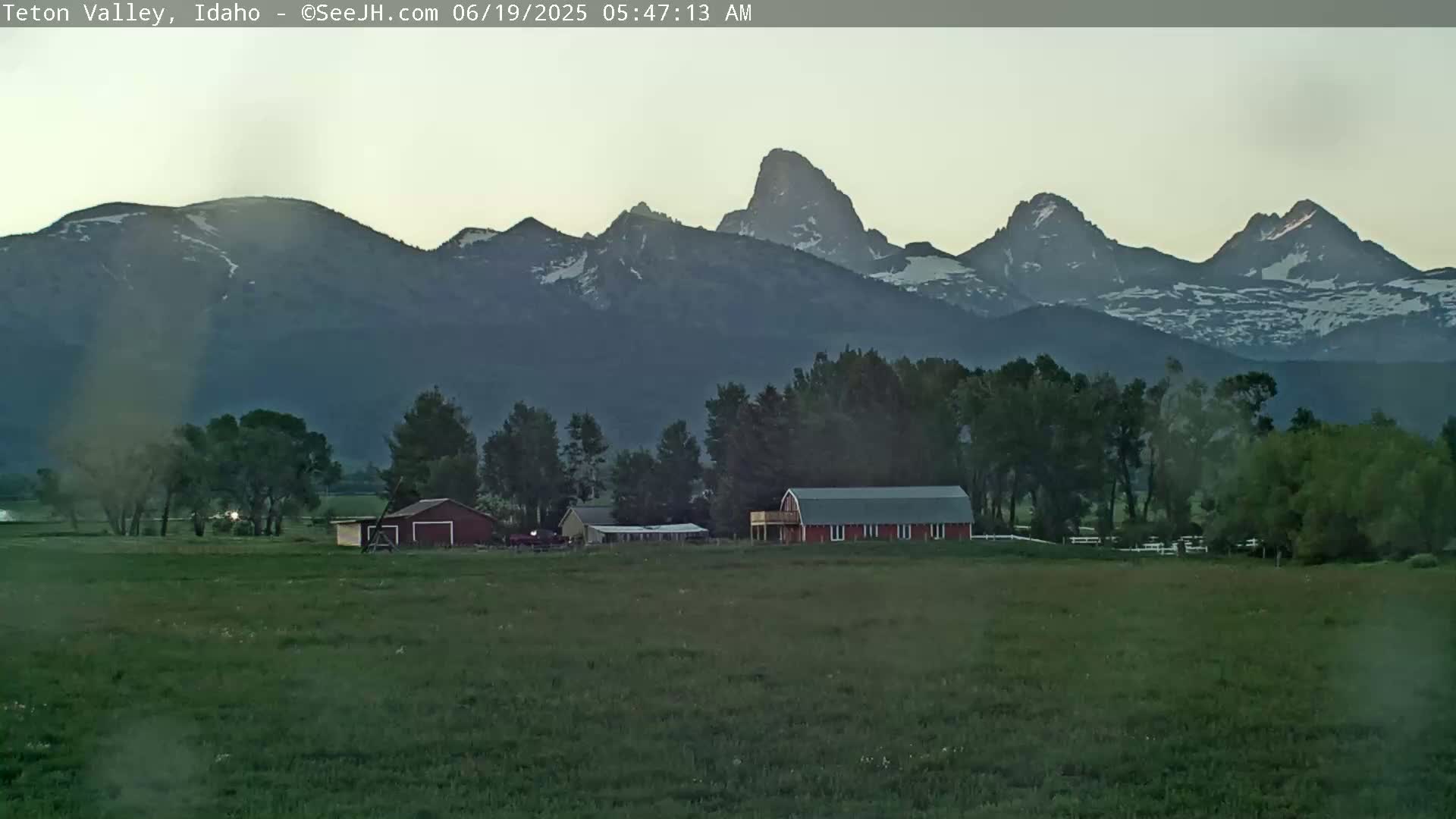 A red barn and other farm buildings sit in a green field, with a range of snow-capped mountains in the background under a twilight sky.