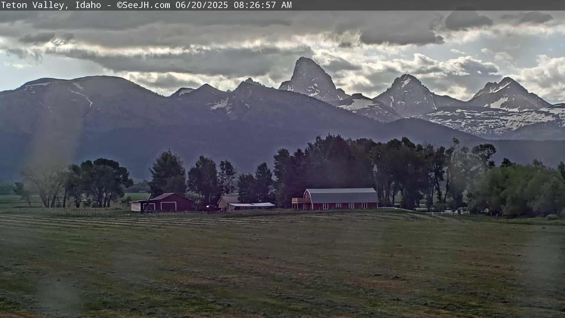 A farm with a red barn sits in a grassy field, overlooked by a range of snow-capped mountains under a cloudy sky.