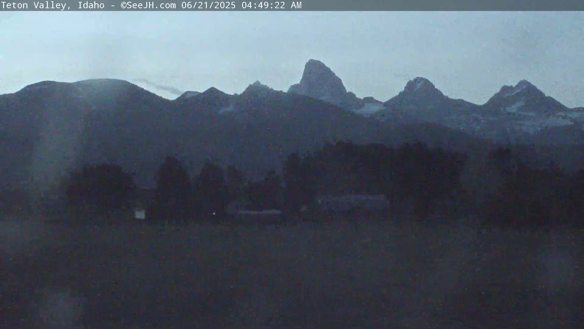 A dark, pre-dawn view shows a range of mountains with some snow on their peaks, behind a dark line of trees and a flat, dark foreground.