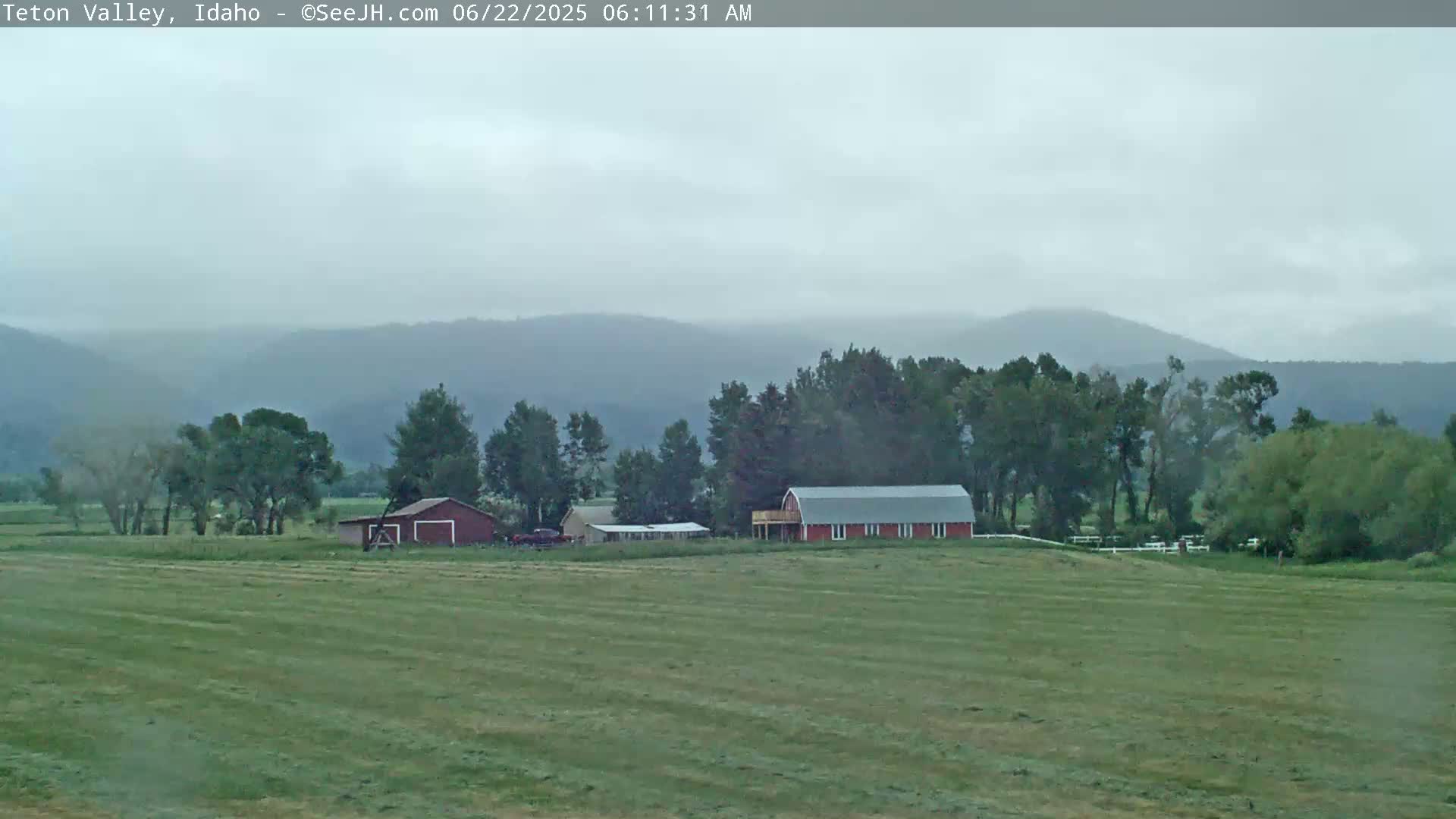 A red barn and other buildings sit on a grassy field, backed by a line of trees and hazy mountains under an overcast sky.