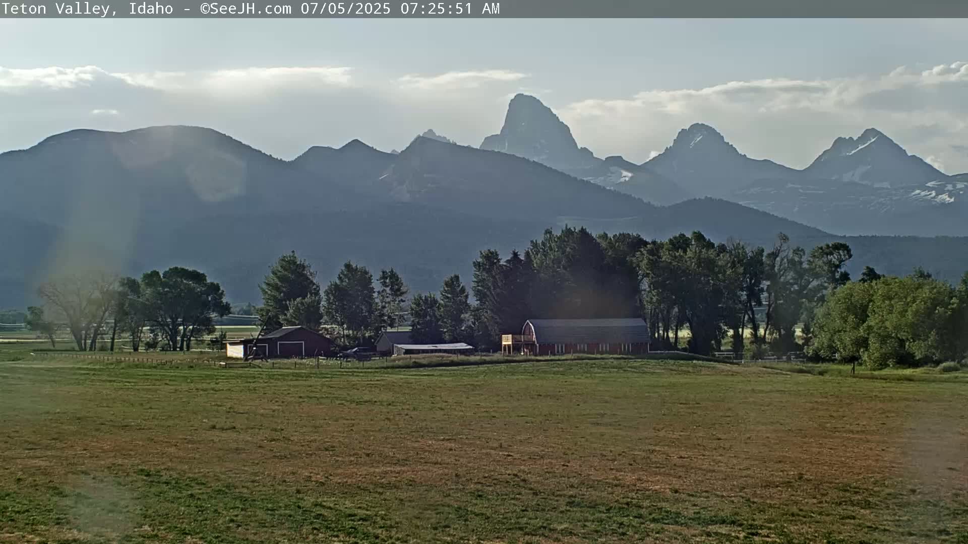 A partly cloudy sky overlooks a flat grassy field with a red barn and other farm buildings, all set against a backdrop of distant, hazy mountains.