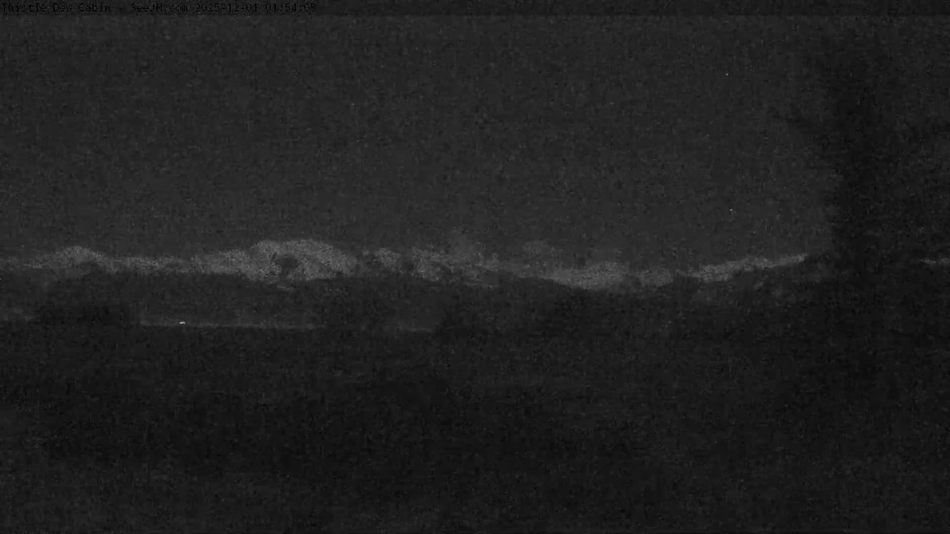 A dark nighttime view displays a distant, faintly illuminated, snow-capped mountain range under a clear sky, with a few small lights visible in the mid-ground.