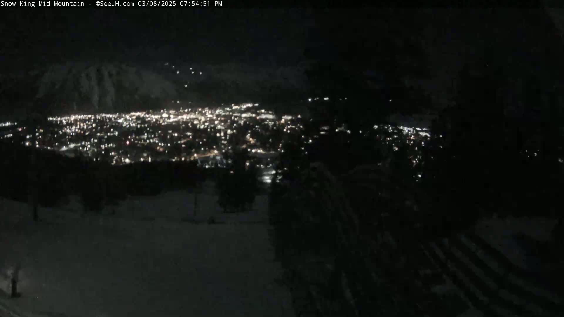 A nighttime view shows a city illuminated below a dark mountain, under a clear sky.